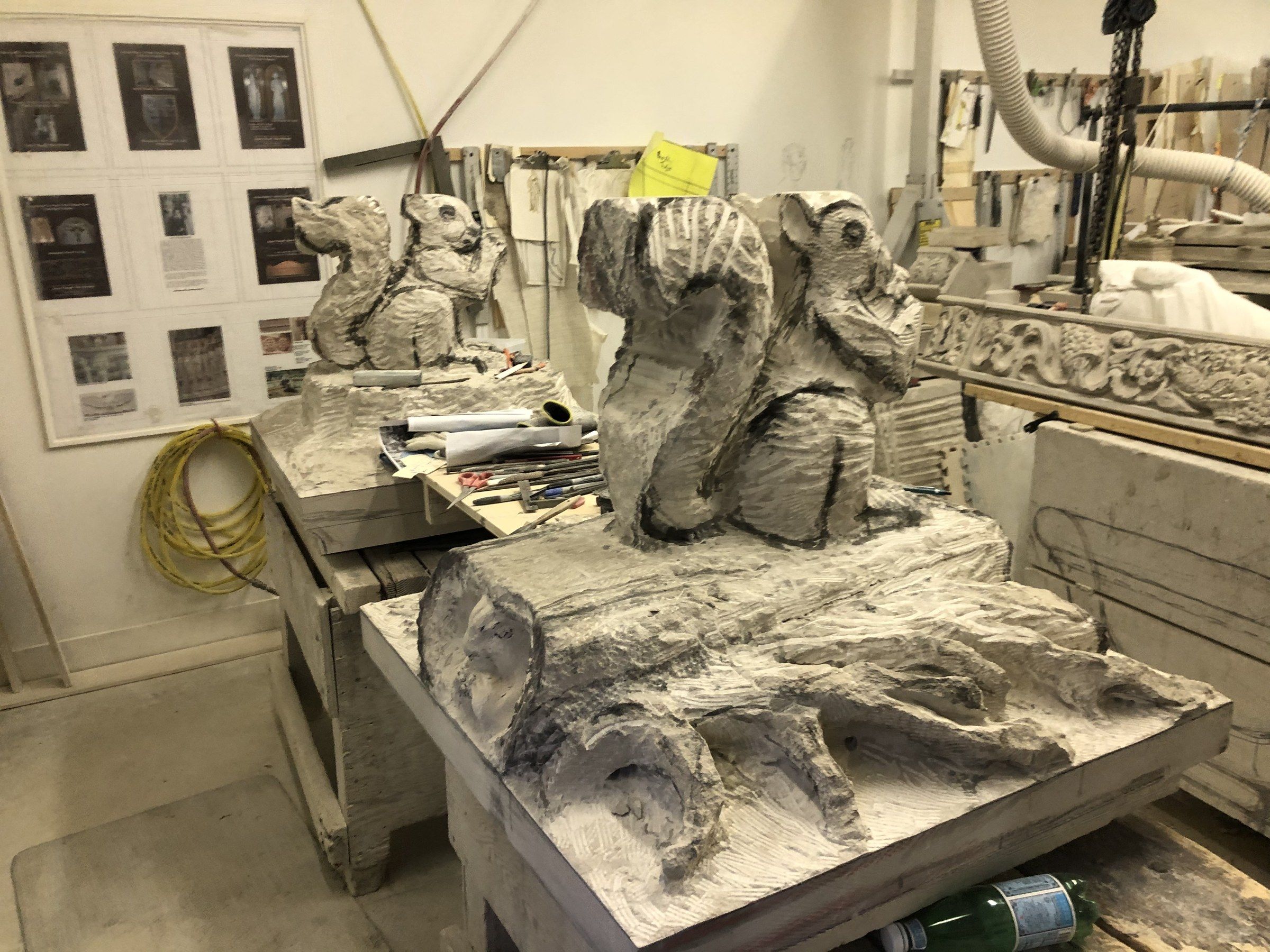 The nearer squirrel taking shape on a carved log, roots spreading across the base; the second pier cap stands on another bench behind.