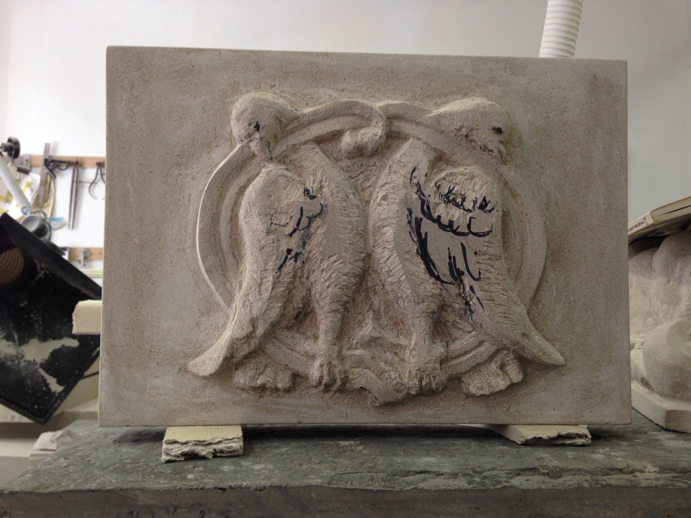 A rectangular limestone relief of two birds standing side by side beneath an interlaced band, inside a round frame; guide lines are still drawn on the wings.