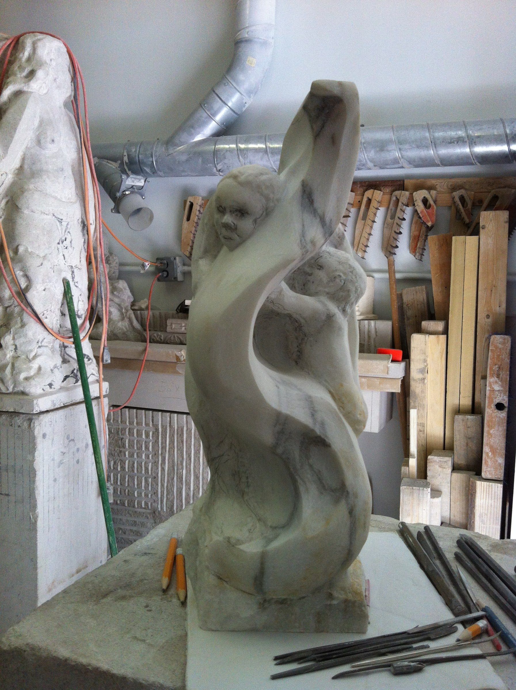 The sculpture risen into a tall twisting form, a child's face near the top and a second figure curled against it, with chisels on the bench.