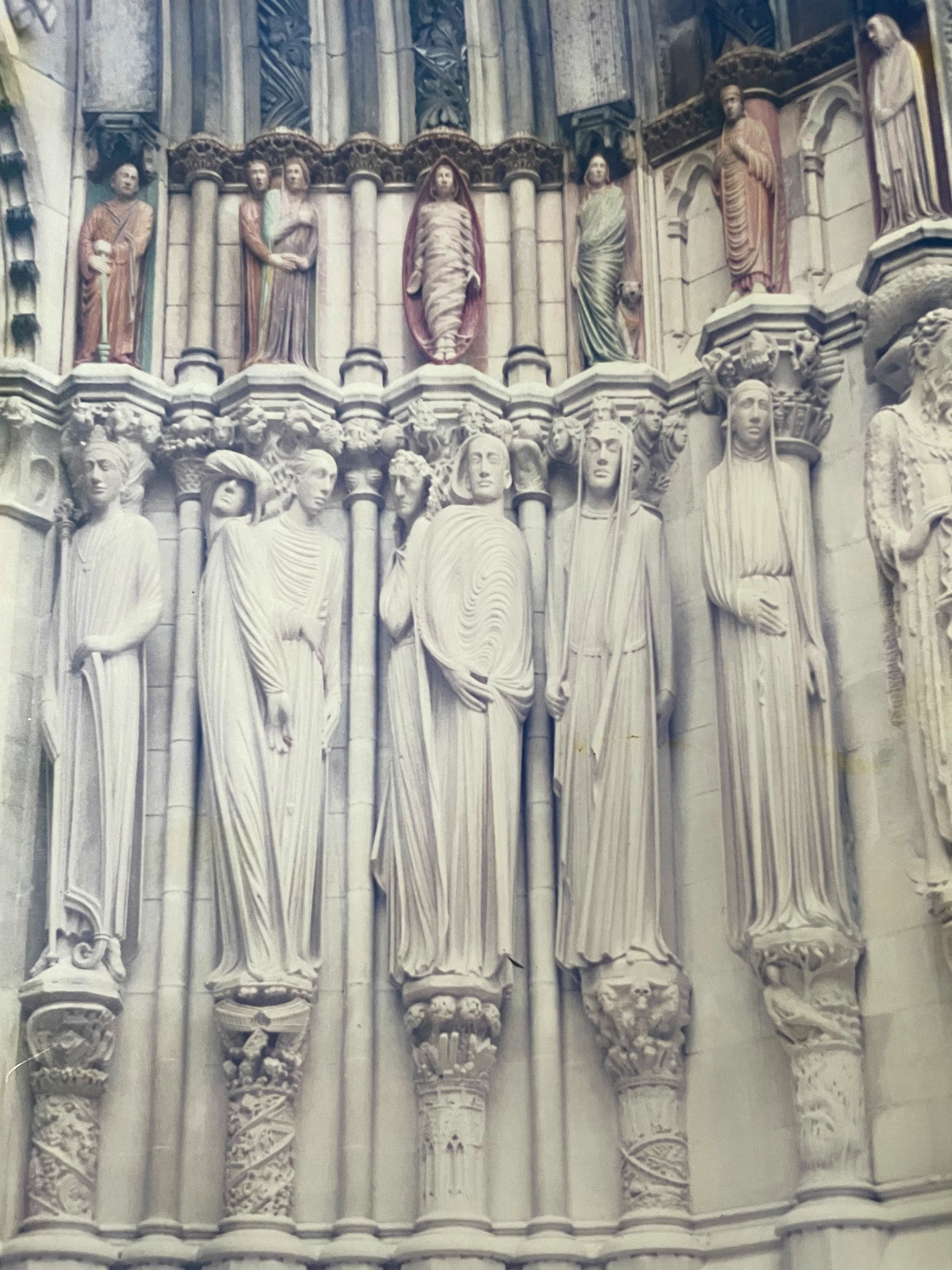 Rows of life-size carved limestone saints standing in the gothic stone portal of the Cathedral of St. John the Divine in New York, beneath an arcade of painted polychrome figures.