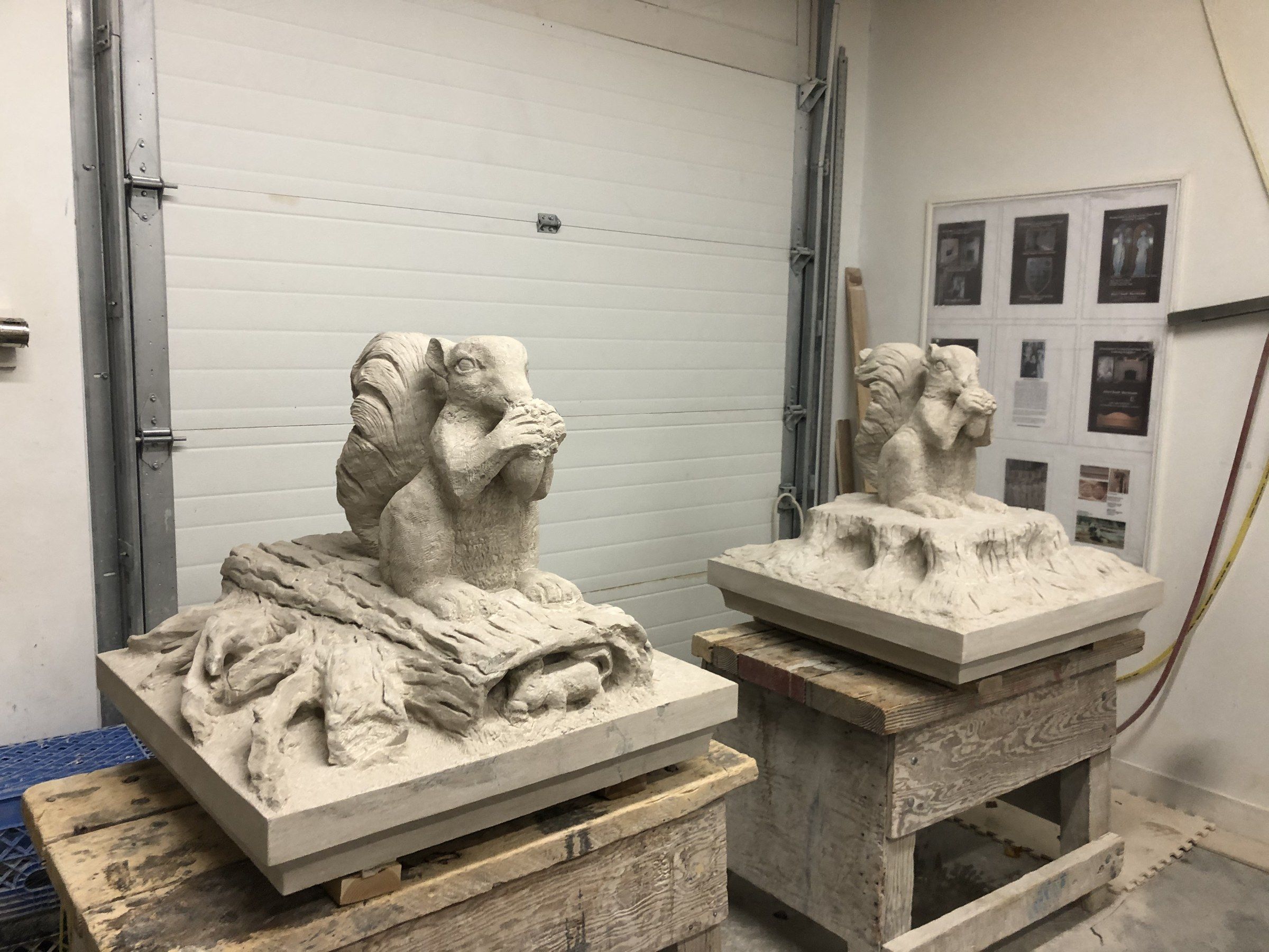 The two finished pier caps side by side in the studio, each squirrel on its carved base, set on wooden stands in front of a white roll-up door.