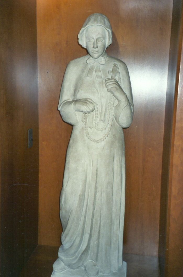 A standing limestone statue of Saint Elizabeth Ann Seton in a wood-paneled niche: she wears a close bonnet, a short cape tied at the collar, and a long plain gown, and holds a rosary with a cross in both hands.
