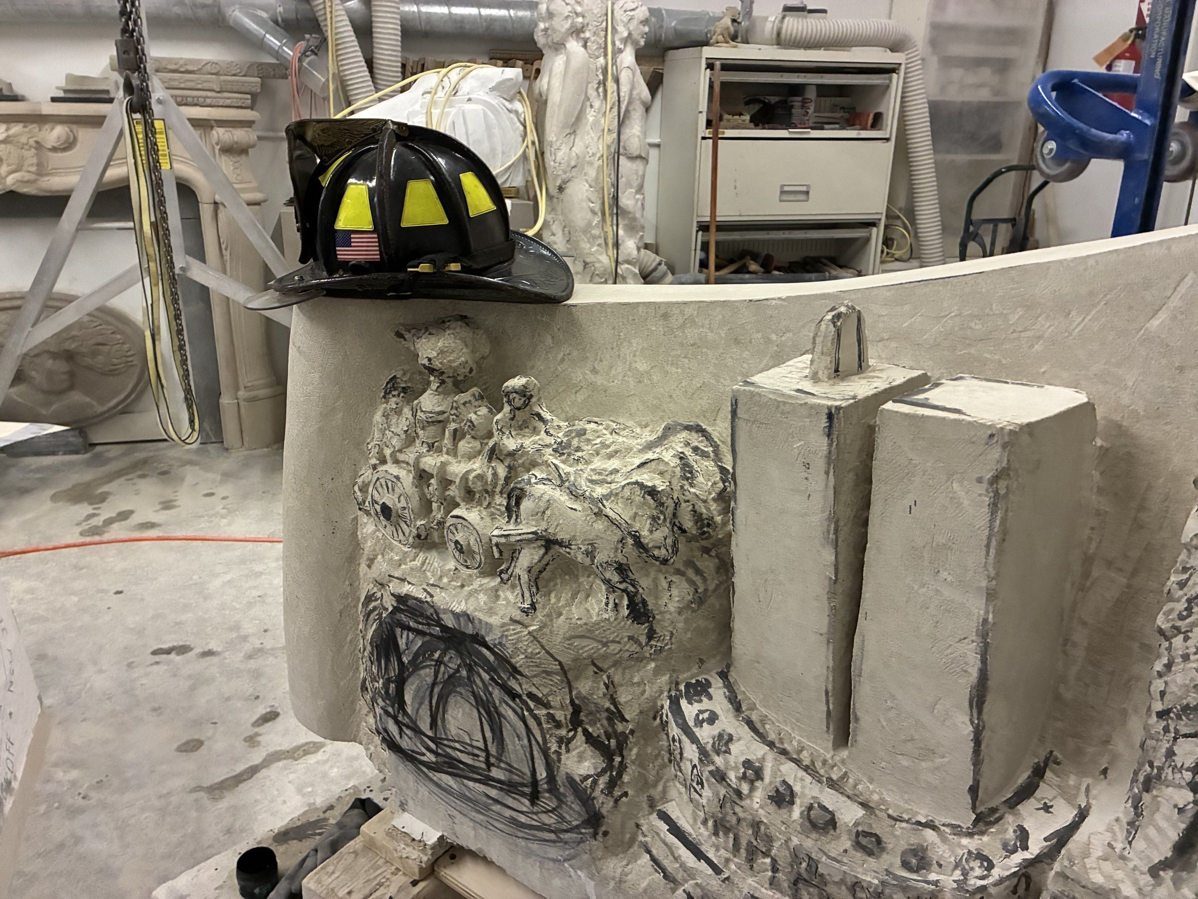A firefighter's helmet, black with yellow reflective bands and an American flag decal, set on top of the stone in June 2026 while the September 11 face is carved: the horse-drawn fire engine roughed out in relief, the Twin Towers still in block form, and the outline of a helmet drawn in black marker below.