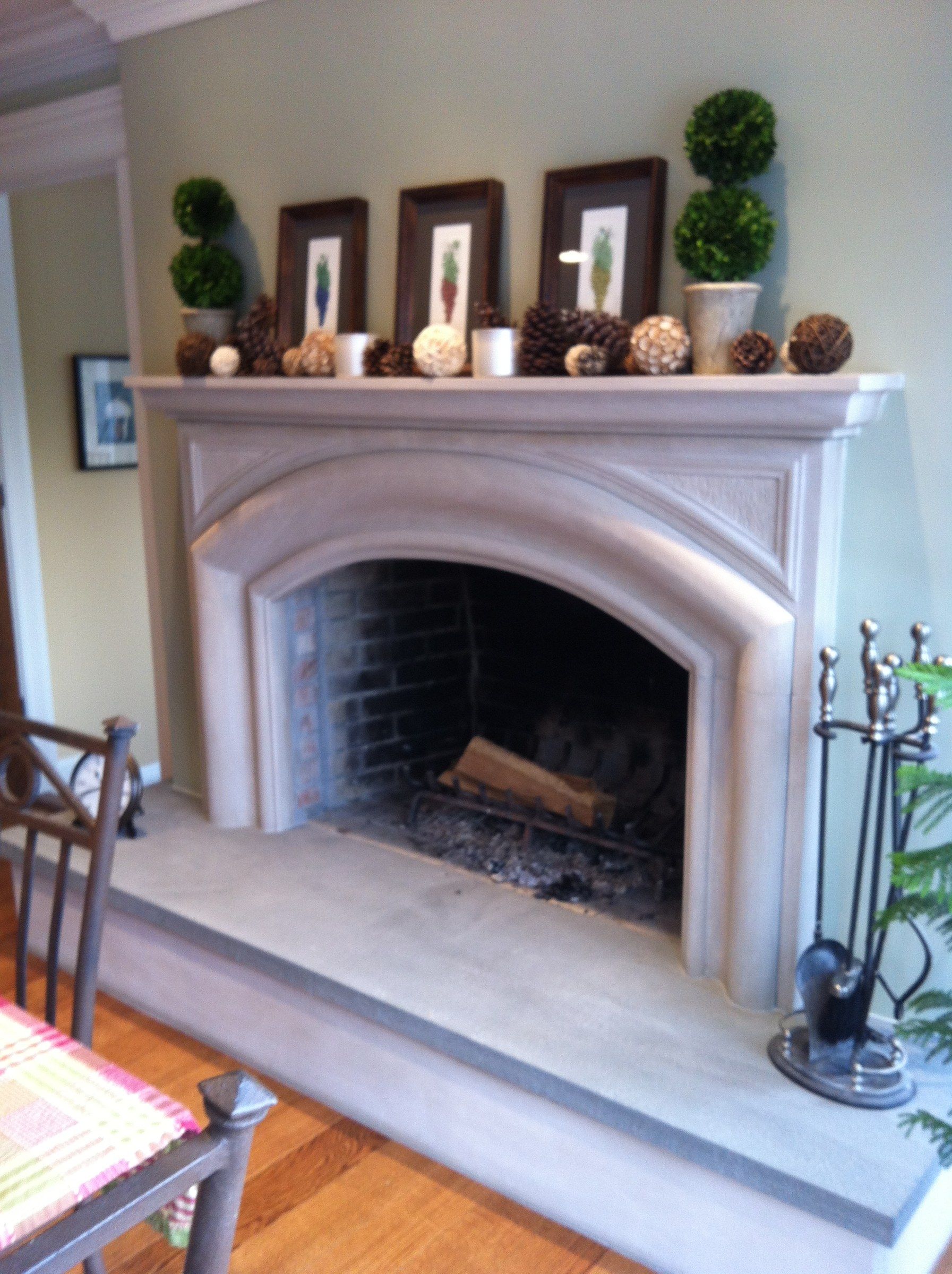 A limestone fireplace in a kitchen: a broad Tudor-arched opening under a plain mantel shelf, on a raised stone hearth, with framed prints and topiaries on the mantel.