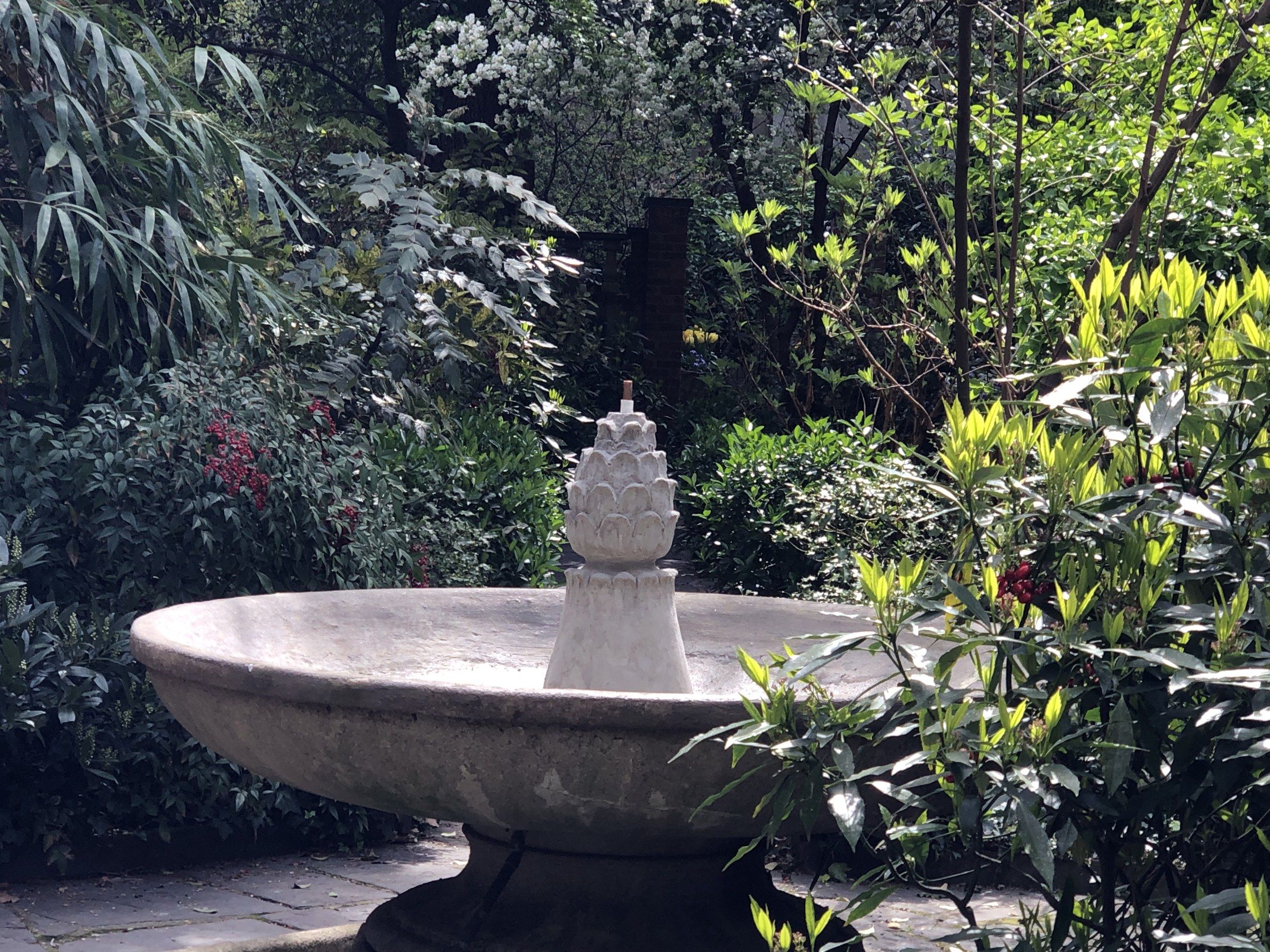 La fontaine vue de biais parmi les plantations, le fleuron s'élevant au centre de la vasque.