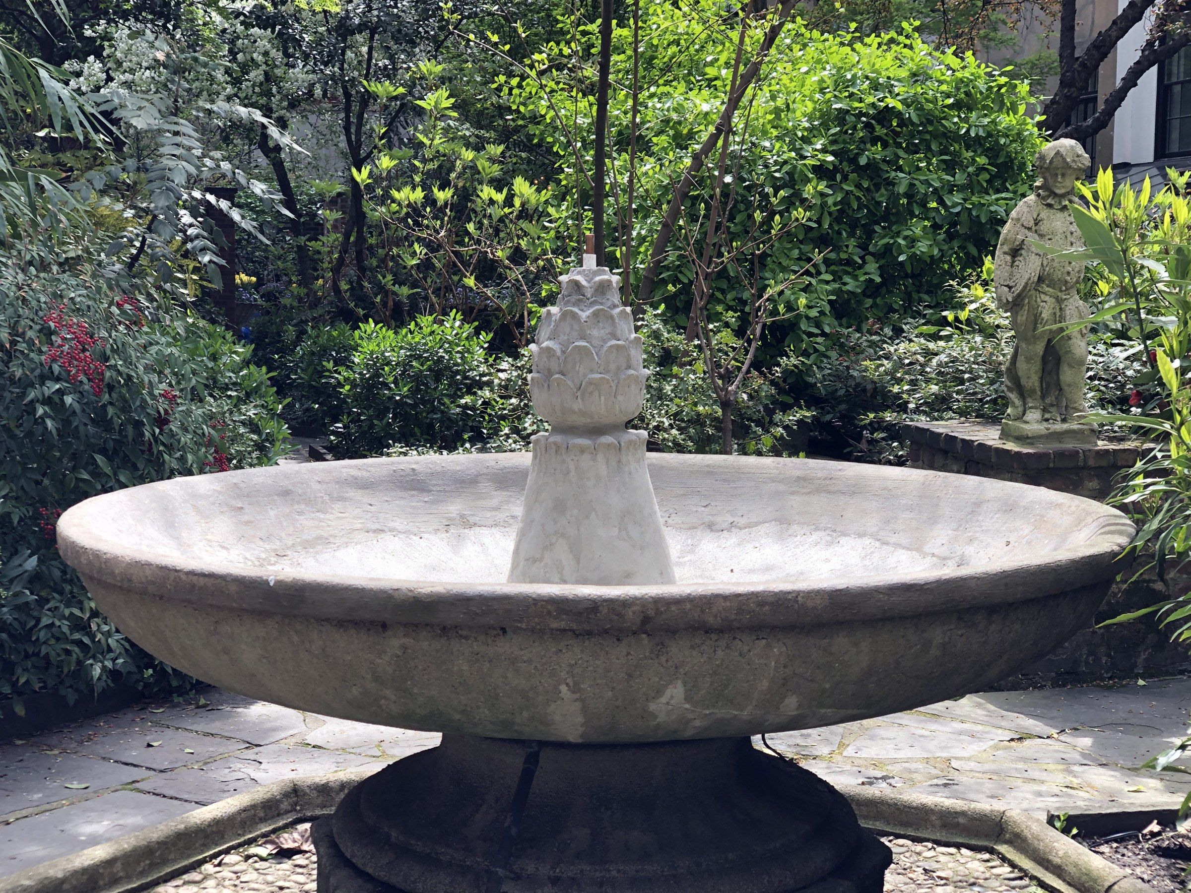 A wide, shallow round fountain basin with a leafy, pine-cone-shaped finial at its center, in a shaded garden with flowering shrubs, paving stones and a small statue of a boy behind.