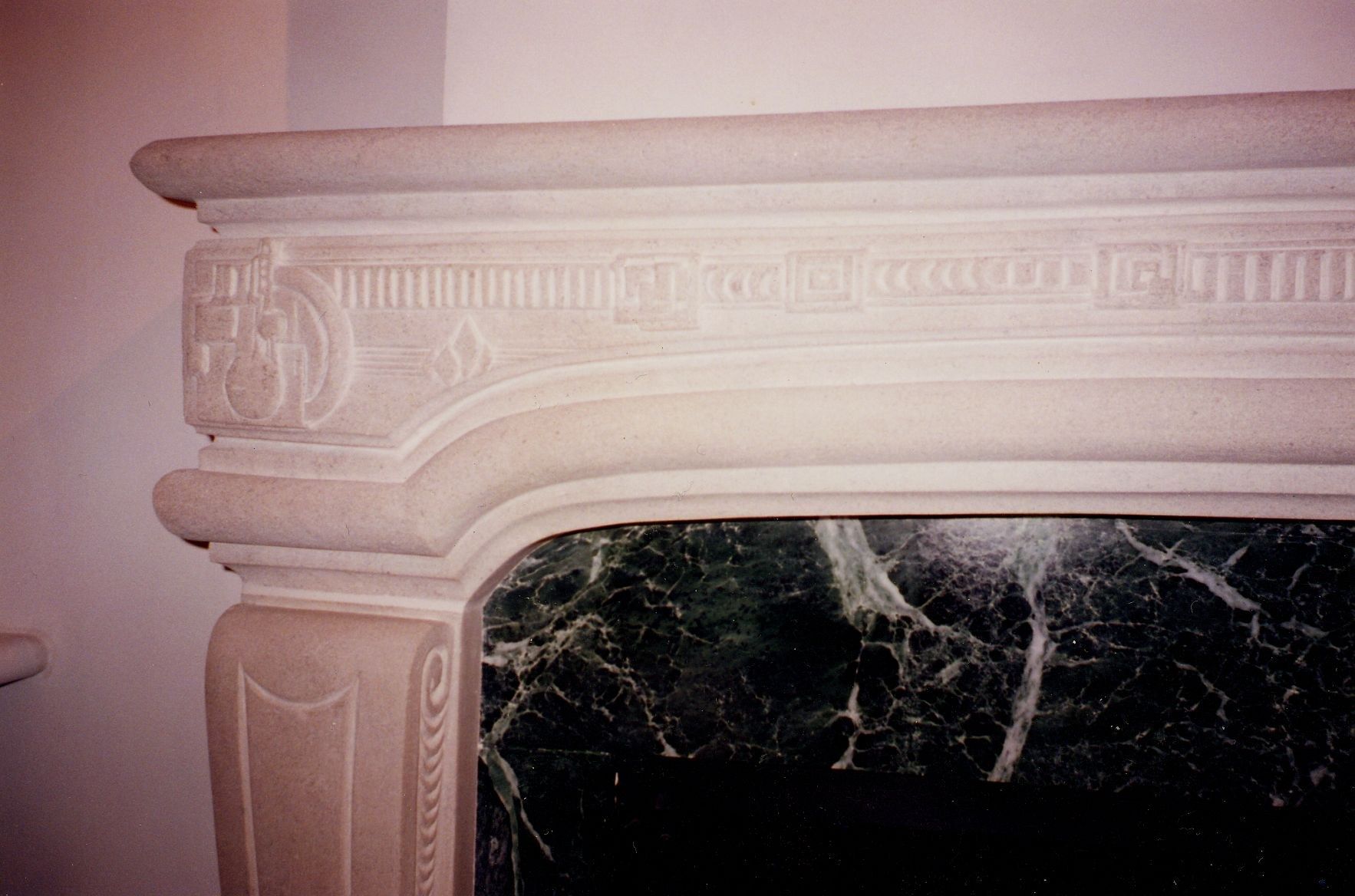 Close-up of the left corner: the rounded shelf, a frieze of fluted and stepped geometric panels, a corner block carved with an abstract geometric motif of mechanical and spatial forms, and the green veined marble slip below the curved opening.