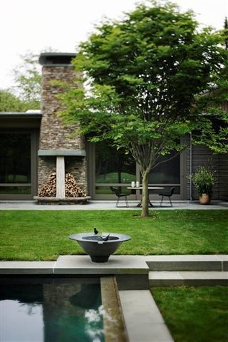 Freestanding outdoor stone fireplace in a landscaped garden on Long Island — walls of stacked natural stone, with a carved limestone column at the hearth, viewed across a lawn with a central tree, boxwood shapes, and a reflecting pool with a sculpted stone basin.