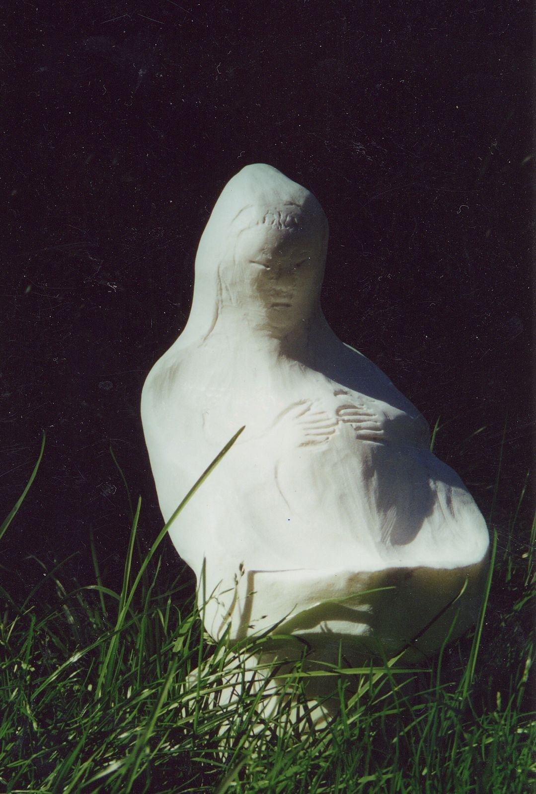 A small white marble figure of the Madonna set in long grass against a dark background: a compact, veiled form with lowered eyes, its features and the hands crossed over the chest drawn in fine incised lines on a smooth, softly polished surface.