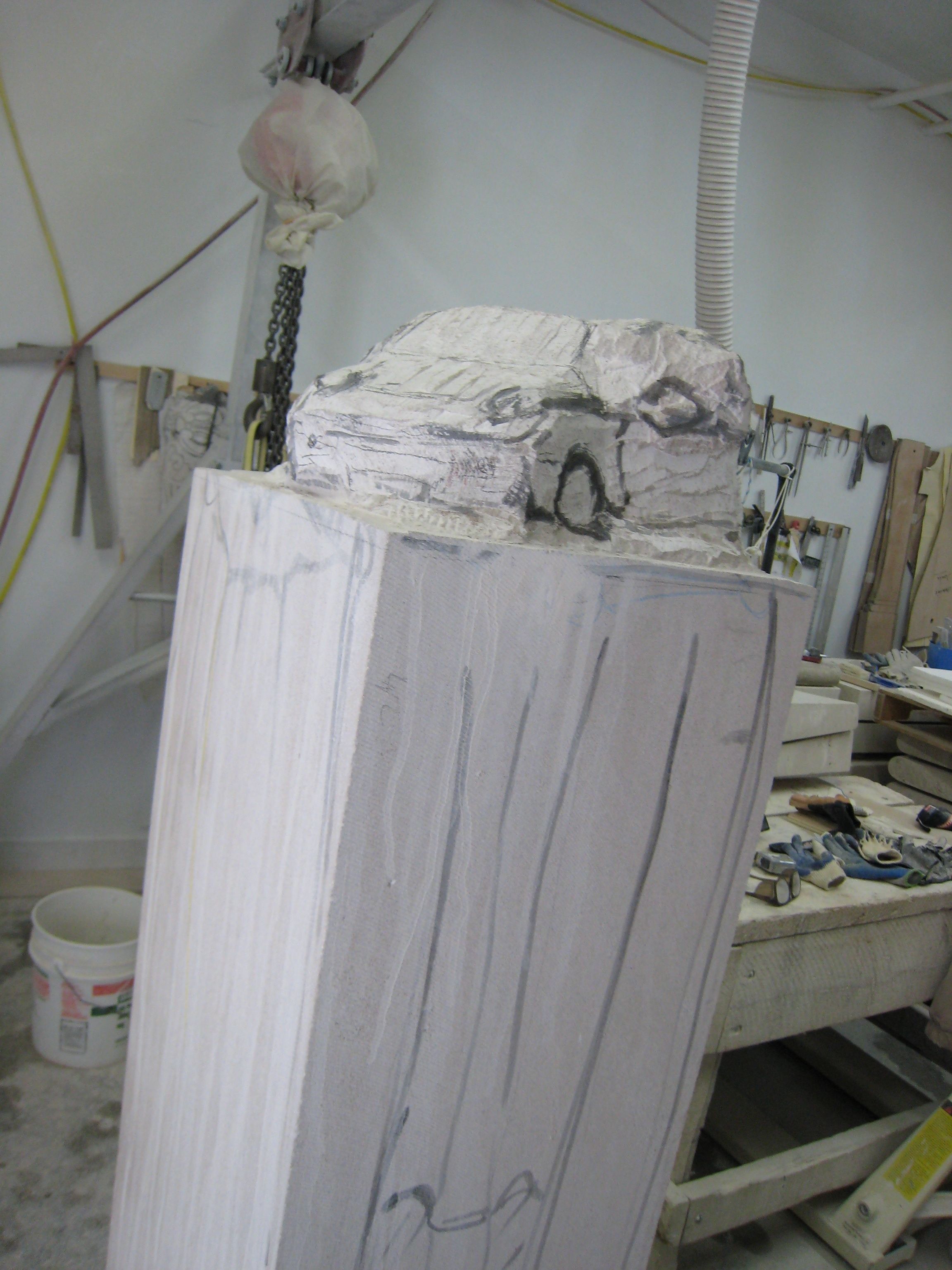 The stone sports car partly roughed out on its pedestal in the workshop, surface still showing chisel marks and pencil layout lines.