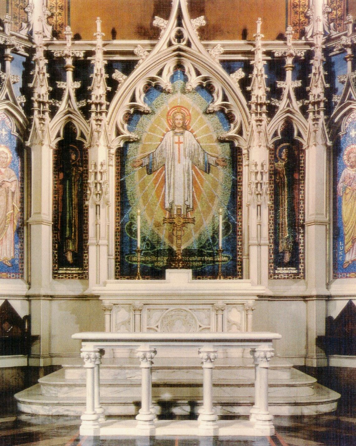 A white marble communion table standing on the marble steps of a chancel: a molded top carried on slender paired colonnettes with carved foliate capitals, set before a gothic reredos of pinnacles and tracery framing a gold-ground mosaic of Christ.