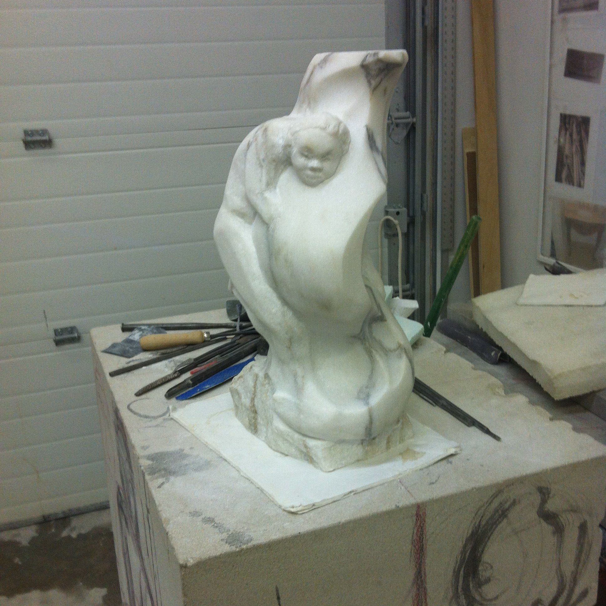 A veined white marble sculpture on a stand in the studio: a child's face emerges near the top of a tall, curving form wrapped around a second figure, with chisels laid at its base.