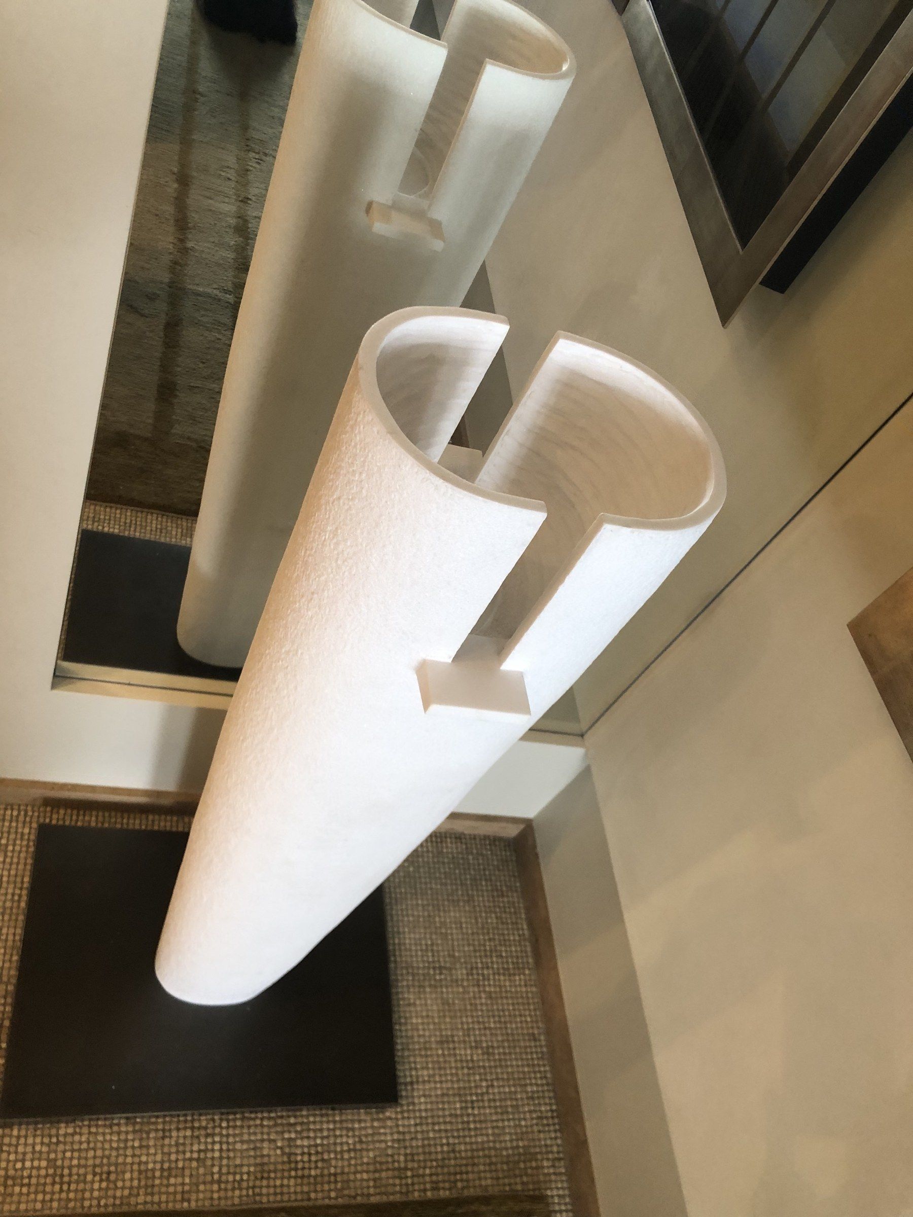 The installed sculpture in April 2025, seen from above at an angle: the hollow, thin-walled cylinder open at the top, a slot running down from the rim, and its reflection in the mirror behind.