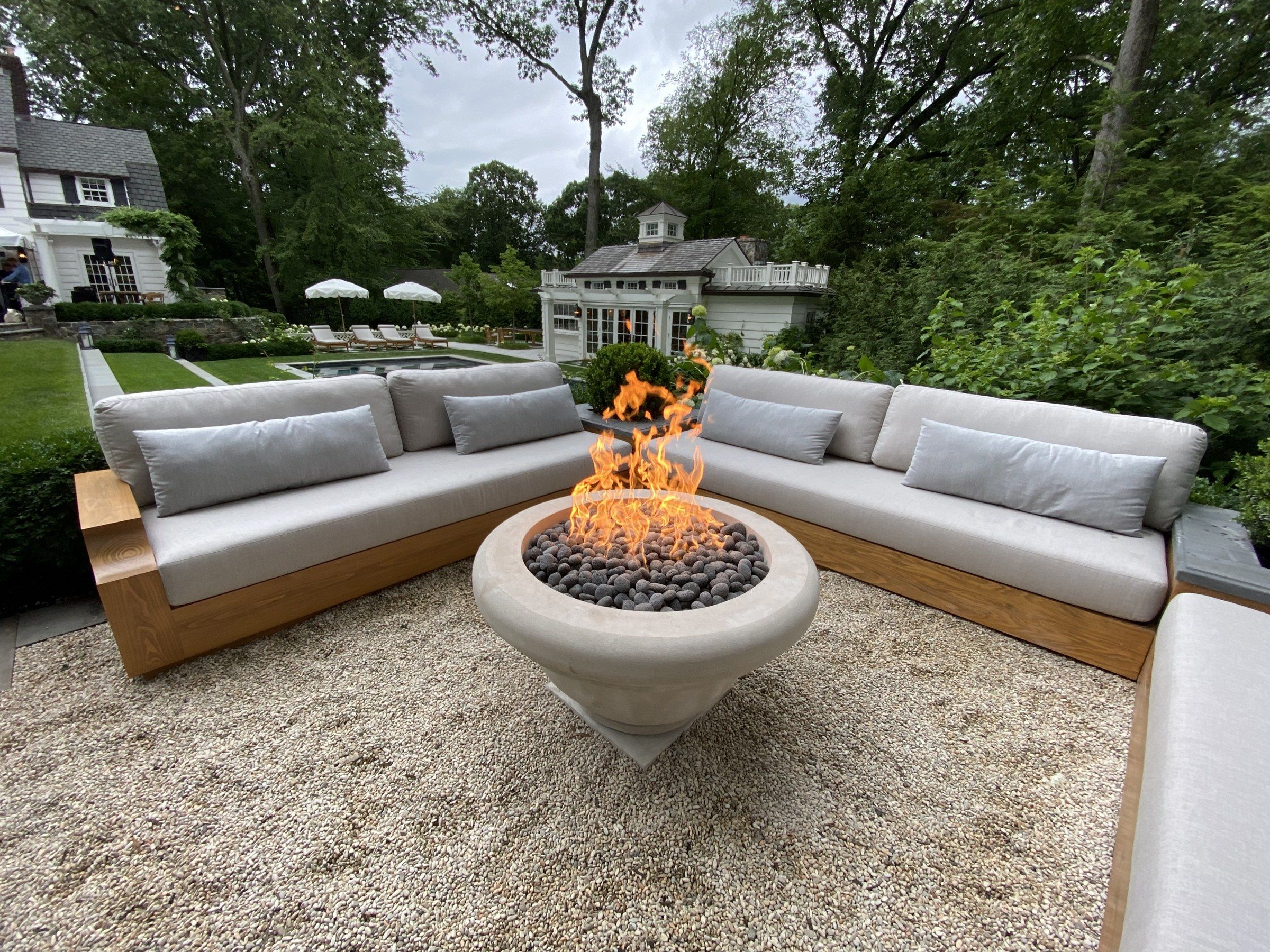 The limestone fire pit in place in a garden: a round bowl with a rolled rim on a round foot and square base, filled with dark stones and a burning fire, surrounded by outdoor sofas on a gravel terrace, with lawn, trees, a pool and houses beyond.