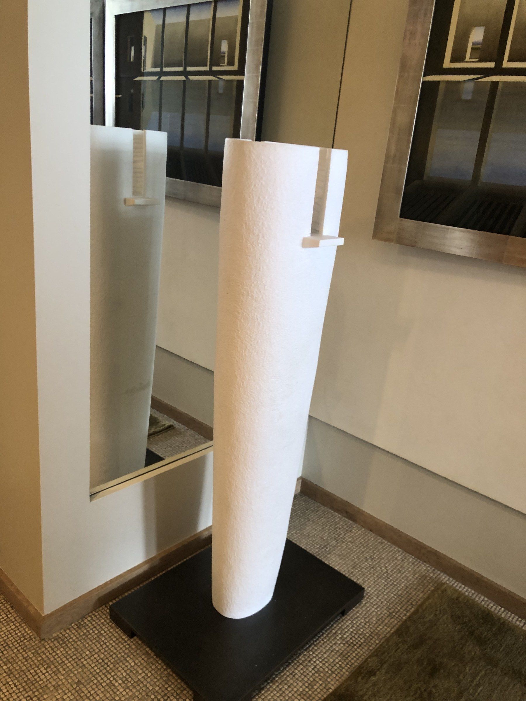 A tall abstract sculpture in white marble standing on a dark square base in a hallway: a slender, slightly tapering cylinder with a lightly textured surface, opened by a narrow vertical slot near the top, with a mirror beside it.