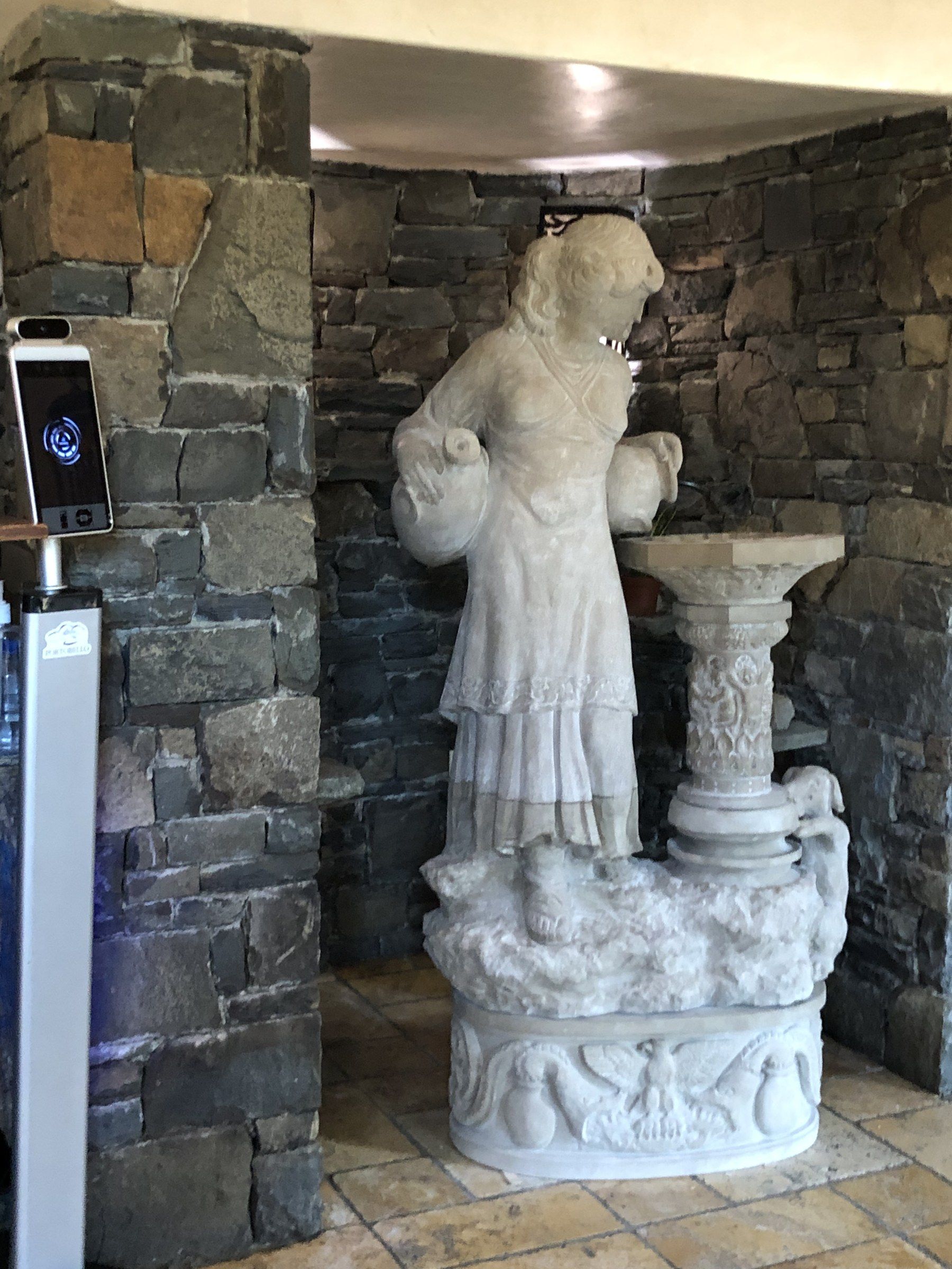 A limestone fountain in a stone-walled alcove: a young woman with a jug under each arm stands on a rocky base beside a column carrying a shallow basin, above an oval pedestal carved with an eagle.