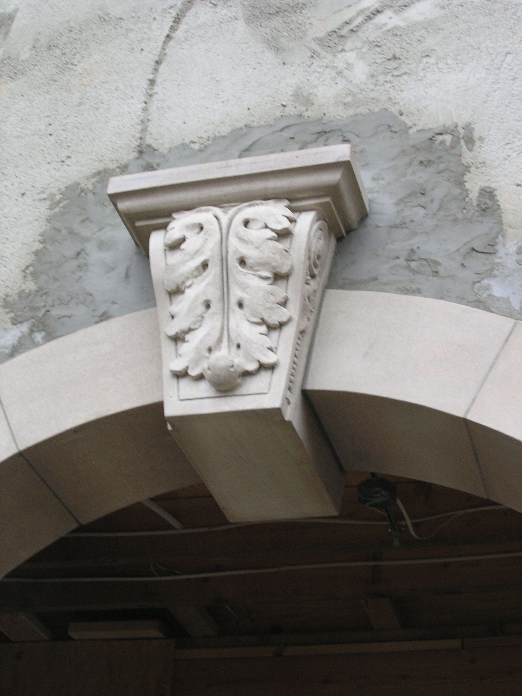 A carved limestone keystone set at the crown of a stone arch on a building under construction: a tapering block with a molded cap, its face carved with an acanthus leaf ending in a small round fruit, and a scroll with a beaded edge along its side.