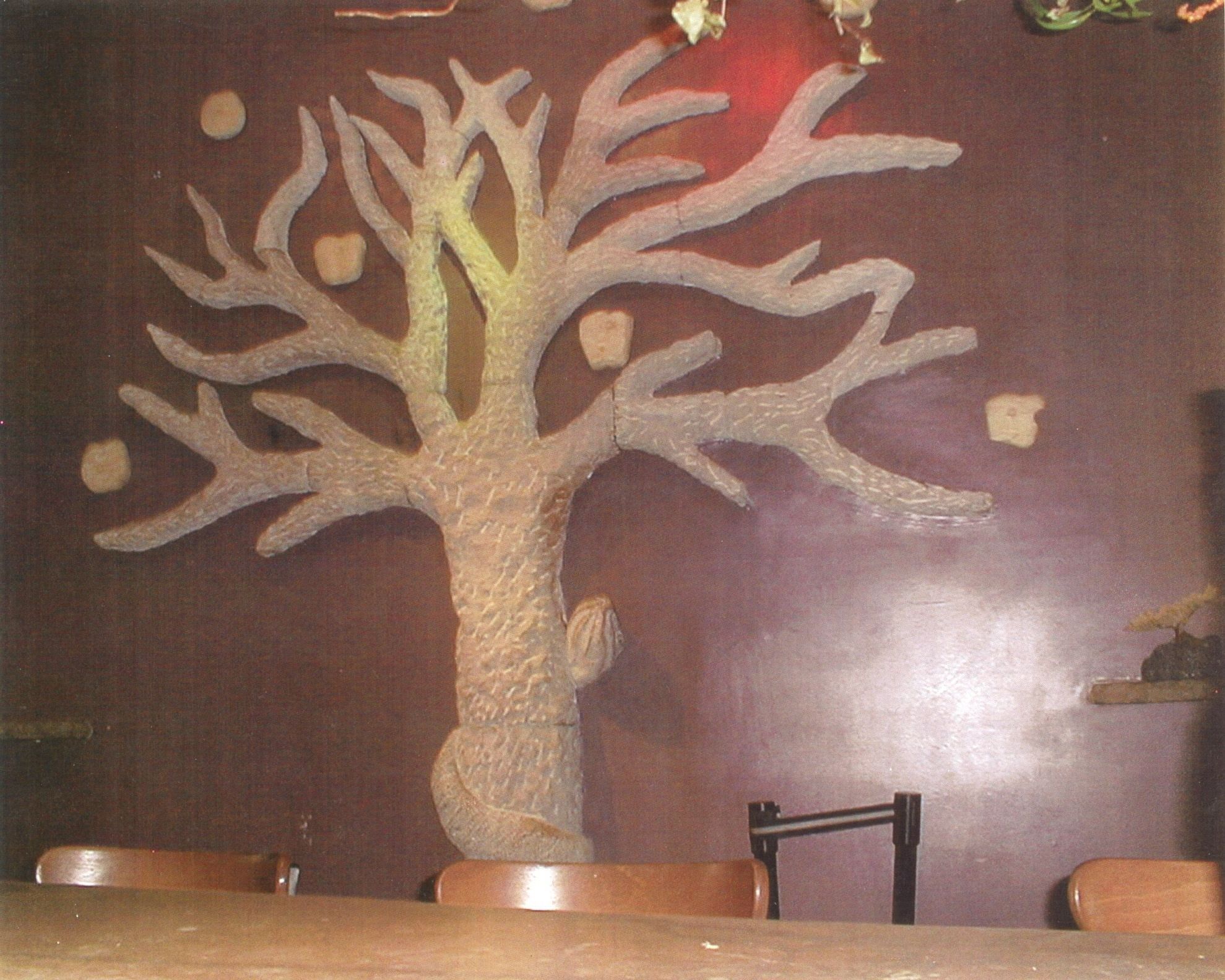 A carved stone tree mounted against a dark wall: a thick trunk with deeply textured bark spreads into bare, forking branches, assembled from several carved sections; a serpent coils around the base of the trunk and winds up behind it, its hooded head leaning out from behind the trunk, and separate carved fruits hang in the spaces between the branches.