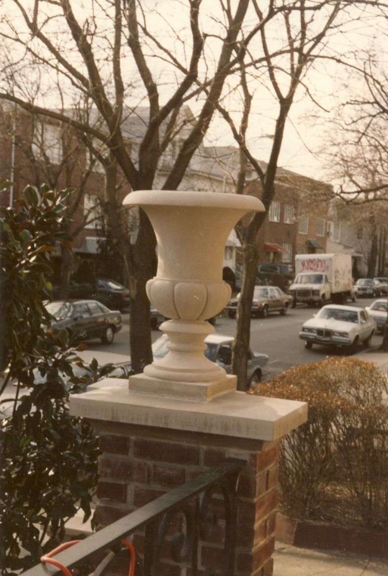 A carved limestone urn of campana form on the capstone of a brick pier at the top of a stoop: a plain flaring bowl over a gadrooned lower body, a turned stem, and a square plinth, with bare trees and a residential street behind.