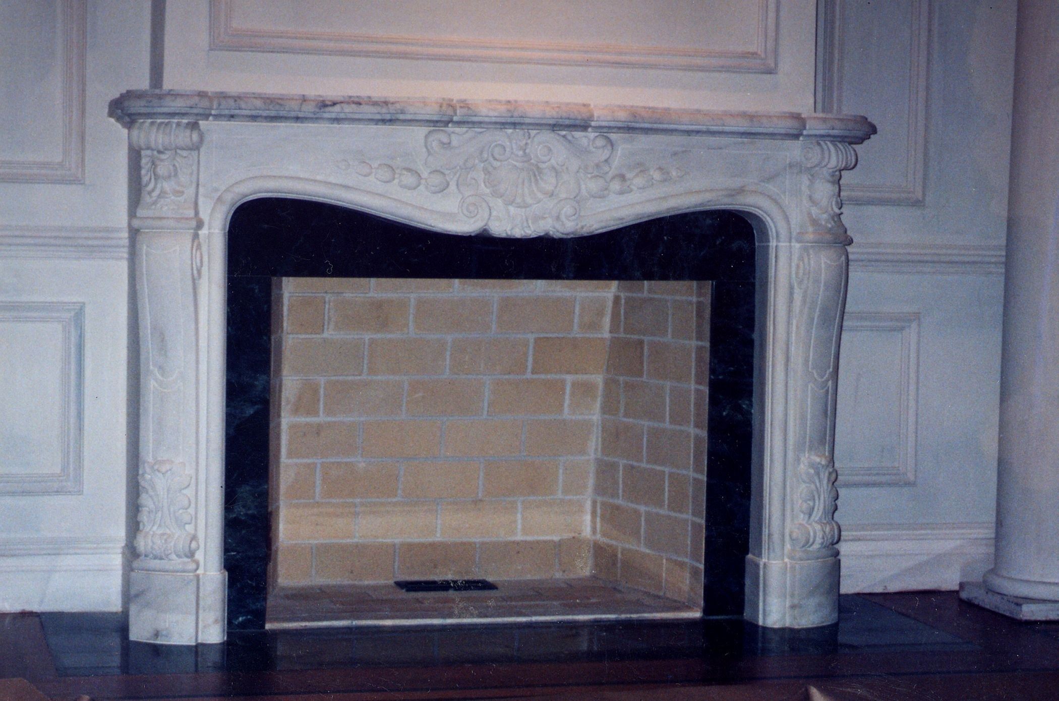 A white veined marble fireplace in the Louis XV manner: a serpentine shelf over a curved frieze carved with a central rocaille shell cartouche and trailing bellflower garlands, cabriole jambs with acanthus scrolls, and a dark green marble slip around a buff brick firebox.