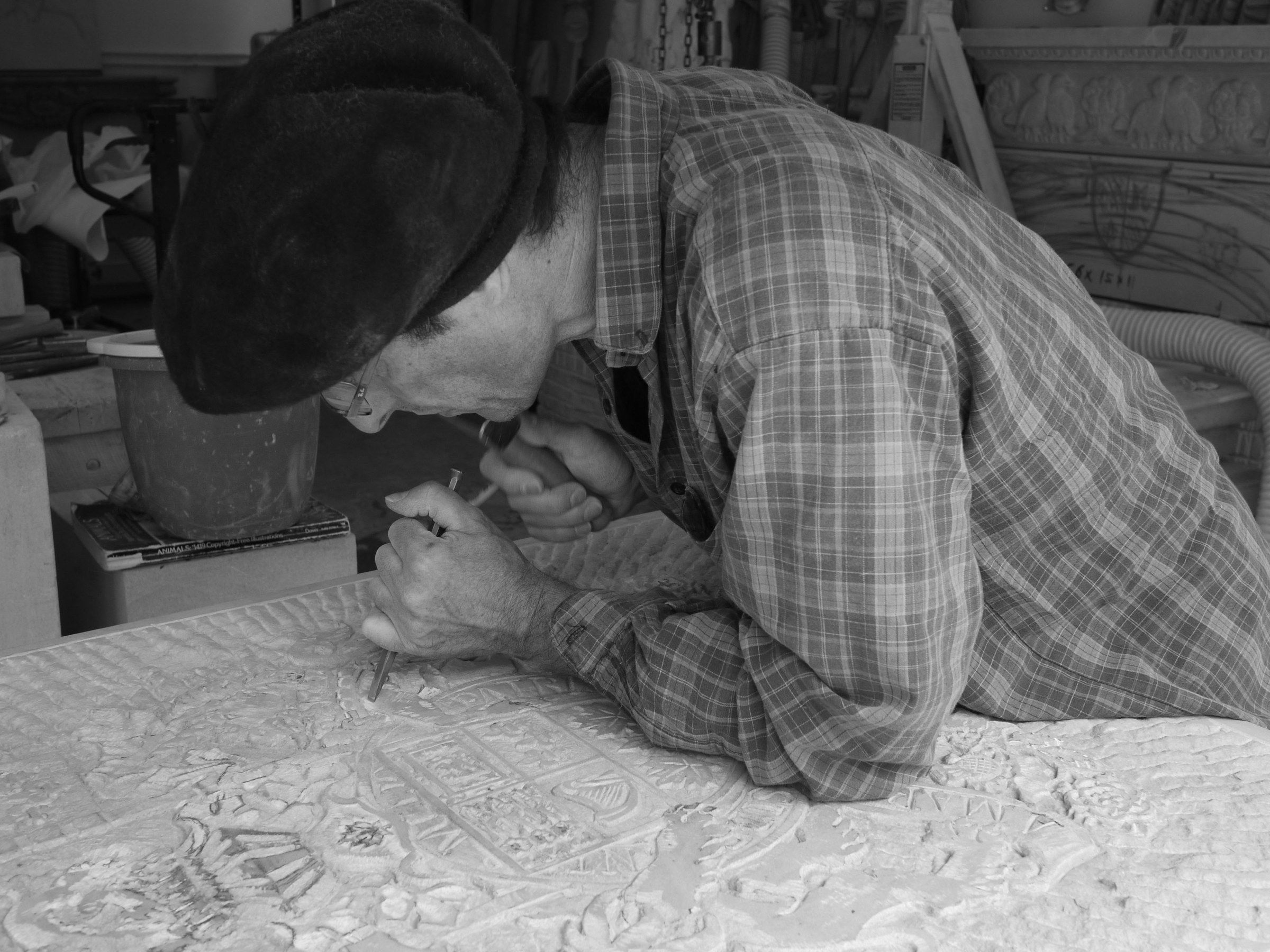 Jean-Claude Marchionni bent over a block of stone in his studio, in a beret and checked shirt, carving a low relief of interlaced panels and rosettes with a chisel. Black-and-white photograph.