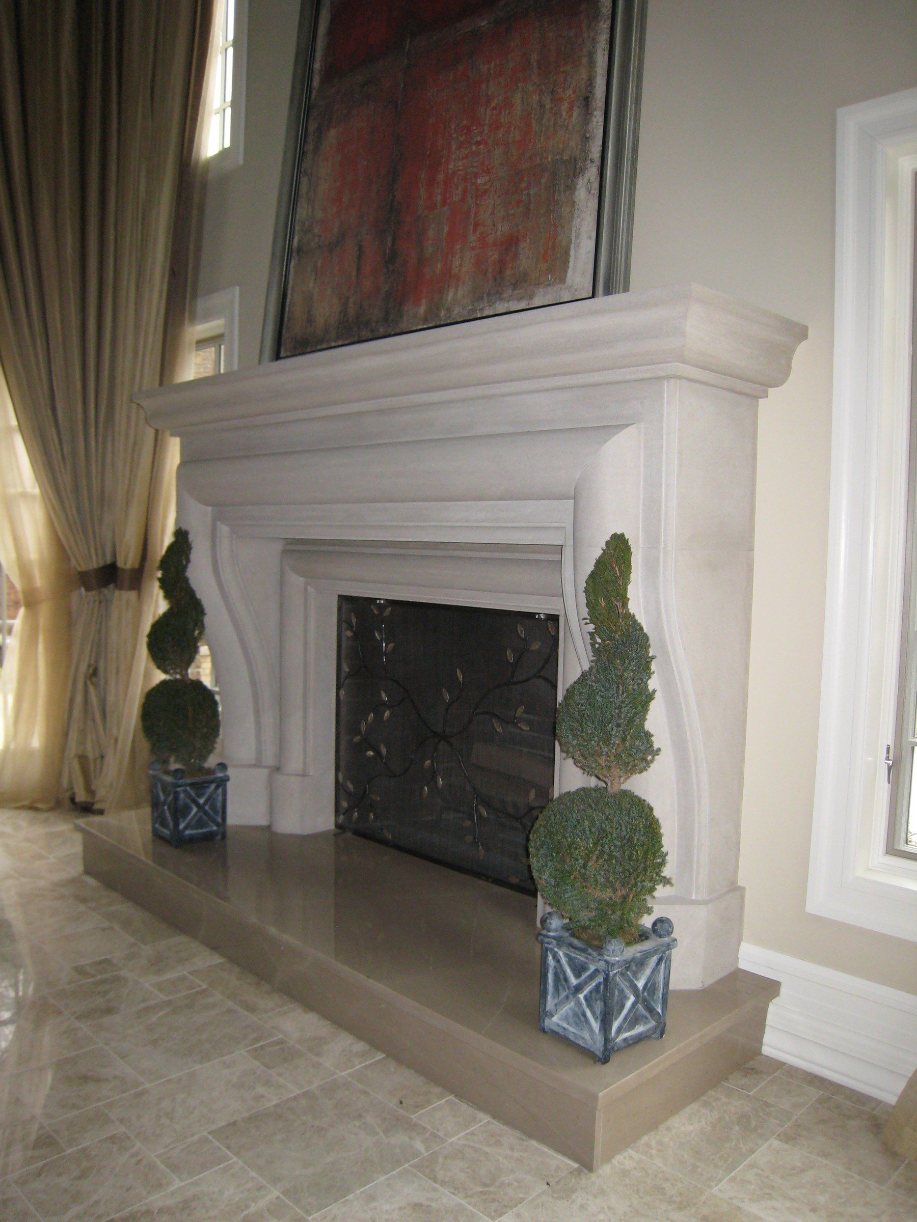 A limestone mantel with a deep bolection profile and curved legs on a raised stone hearth, with a topiary on either side and a large painting above.