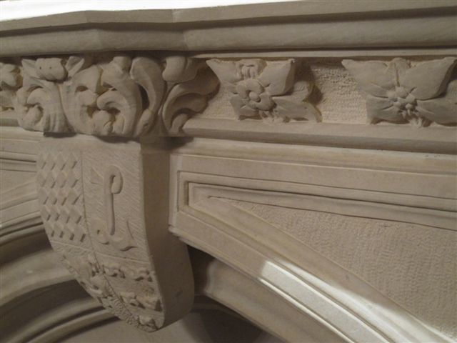 Close-up of the carved mantel: foliate scrolls over the corbels, a row of carved rose medallions set in coffered squares along the frieze, and the central heraldic shield with a carved charge.