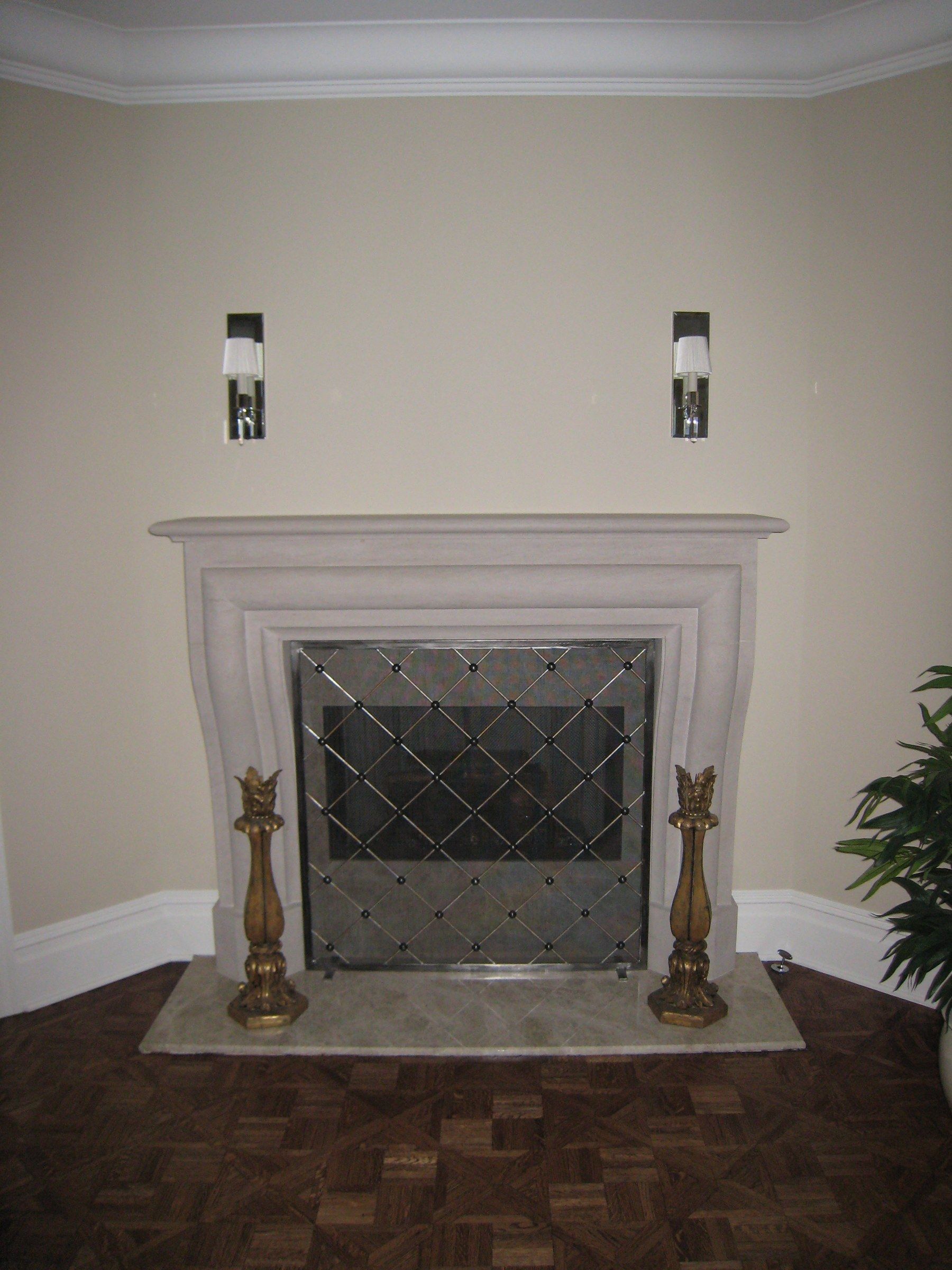 A limestone mantel with a bolection profile seen from the front between two wall sconces, with a lattice fire screen and gilt candlesticks on the hearth.