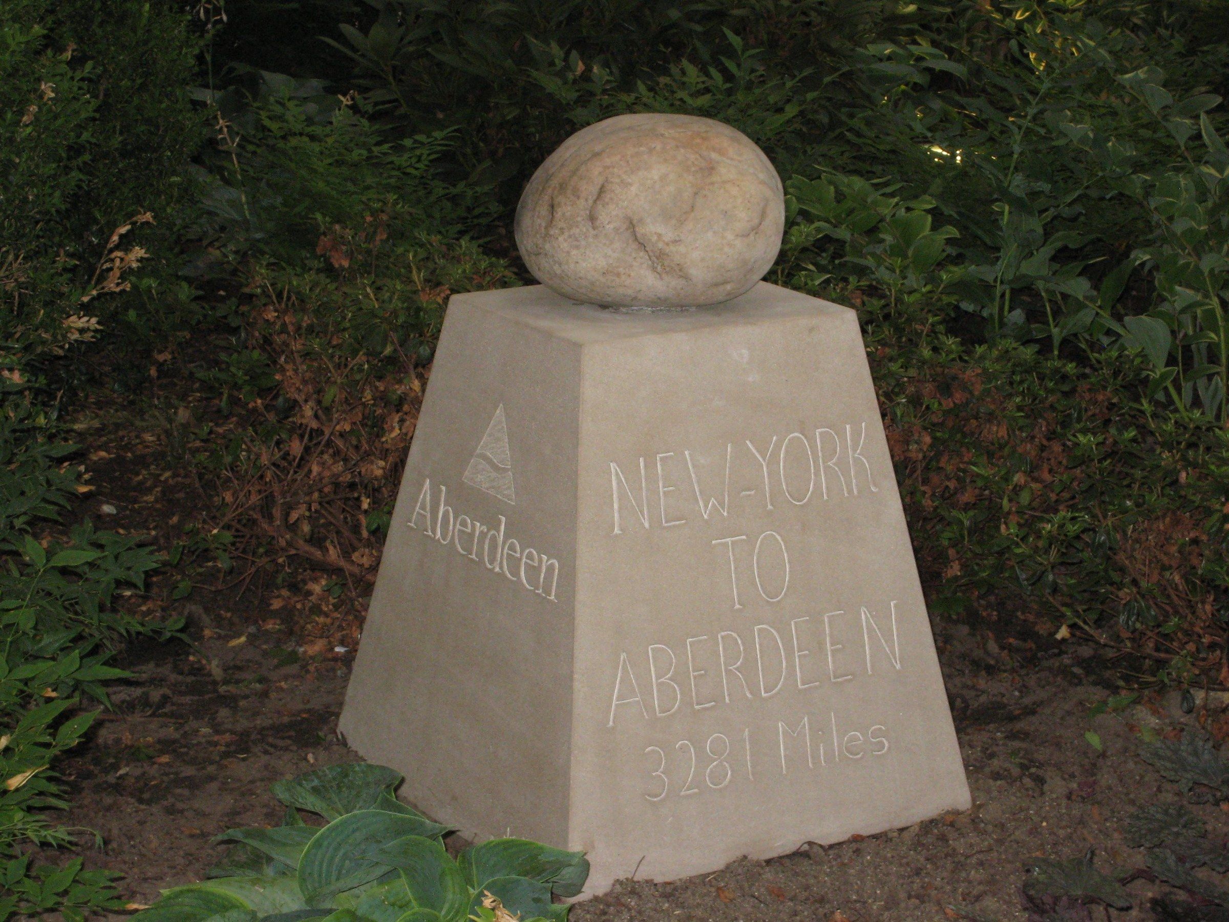 A tapering square limestone marker topped with a rounded boulder, among garden plants, inscribed “NEW-YORK TO ABERDEEN 3281 Miles” on one face and “Aberdeen” beneath a triangular emblem on the other.