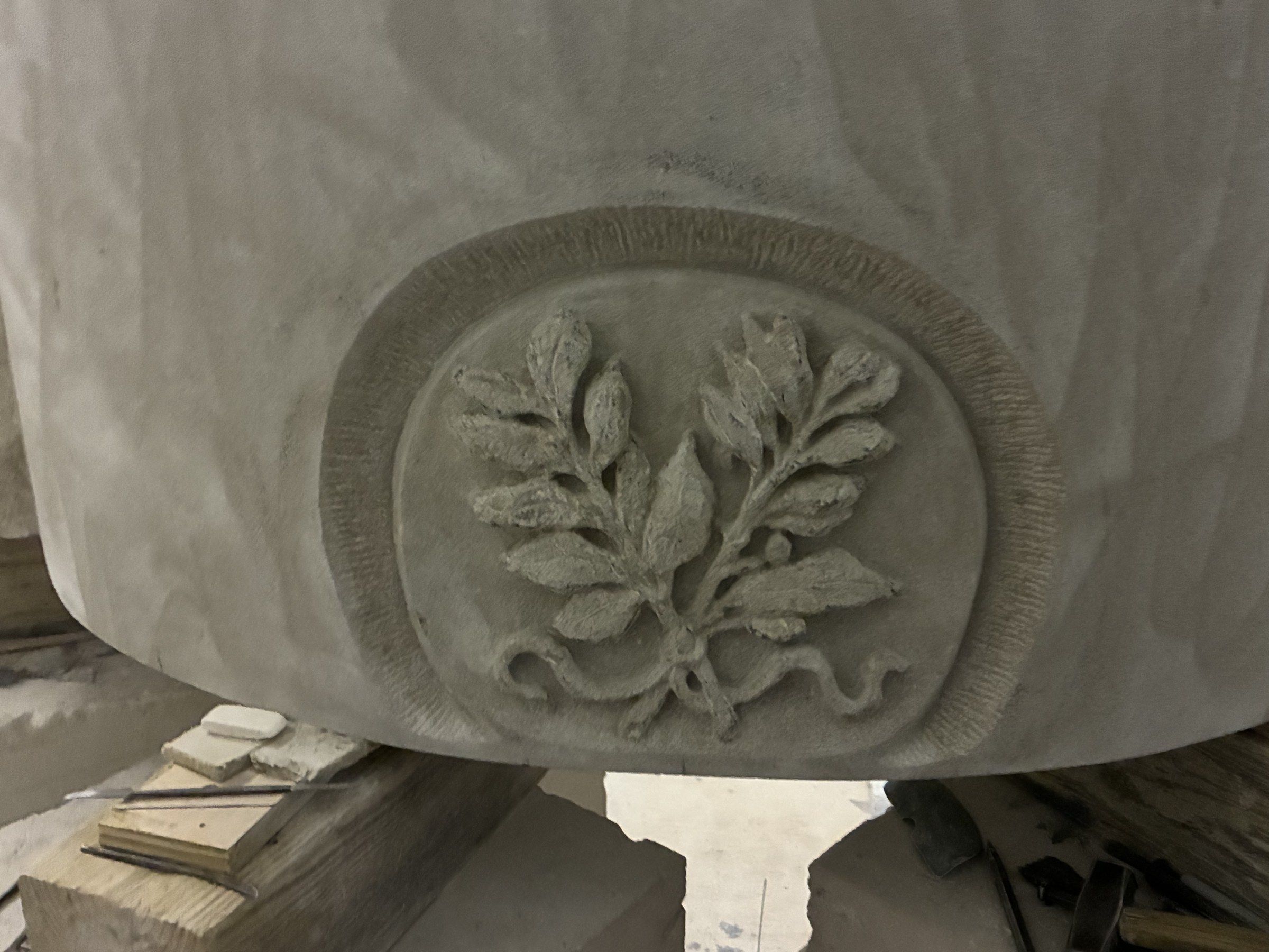 Close-up in August 2026 of an oval medallion carved in low relief with a spray of oak leaves, set into the curved face of the stone.