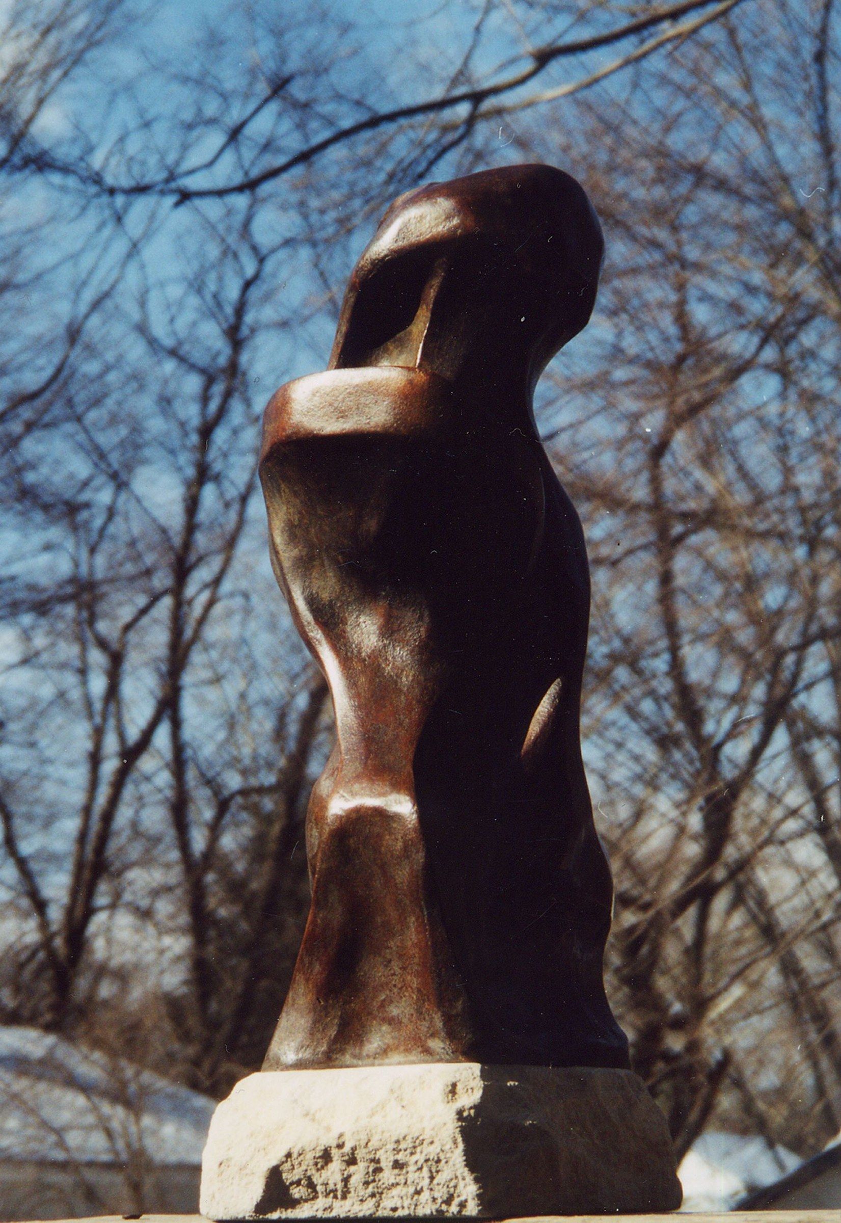 A stylized bronze figure with a warm brown patina, standing on a rough-hewn pale stone base and photographed from below against bare winter branches and a blue sky: a tall, twisting body with a hooded, bird-like head bent forward, pierced by an open loop.