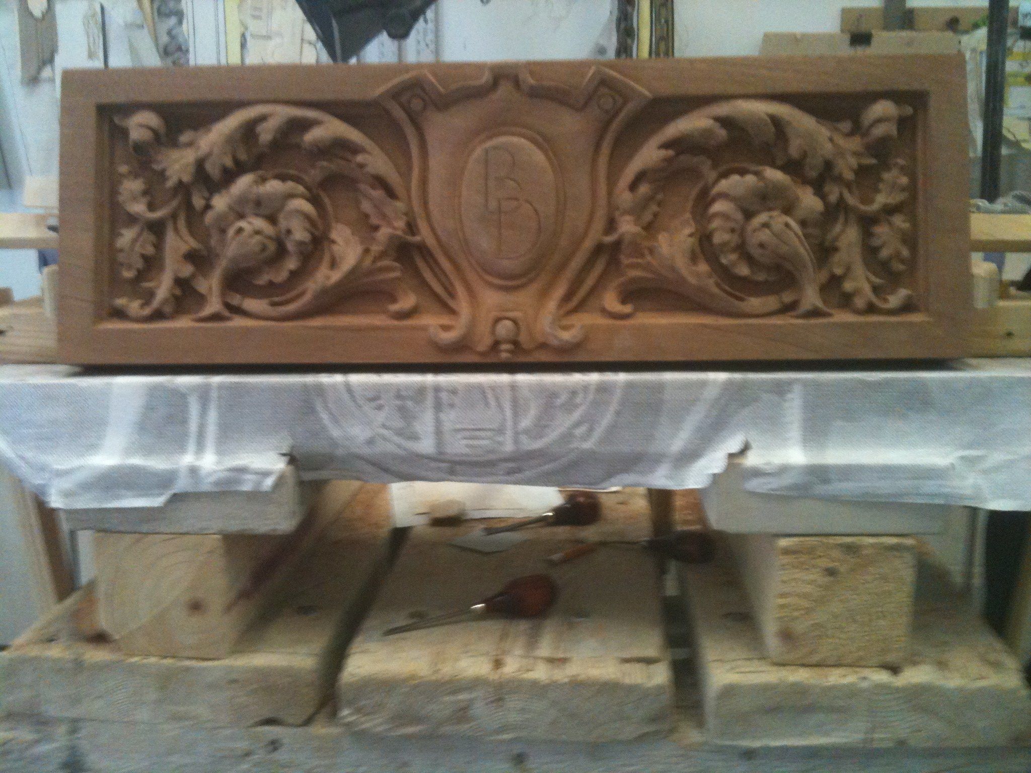 A long carved wood panel on the workbench: a central oval cartouche with a monogram, flanked by scrolling acanthus leaves.