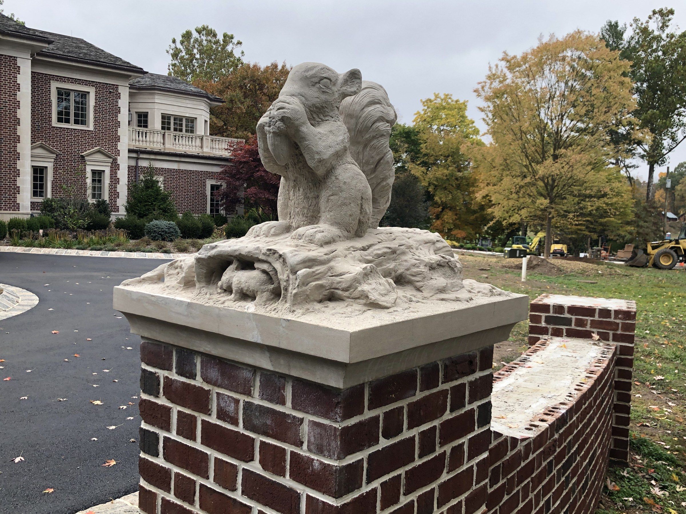 One squirrel seen from the side on its pier, the log and roots beneath it, with the driveway, the house and autumn trees behind.