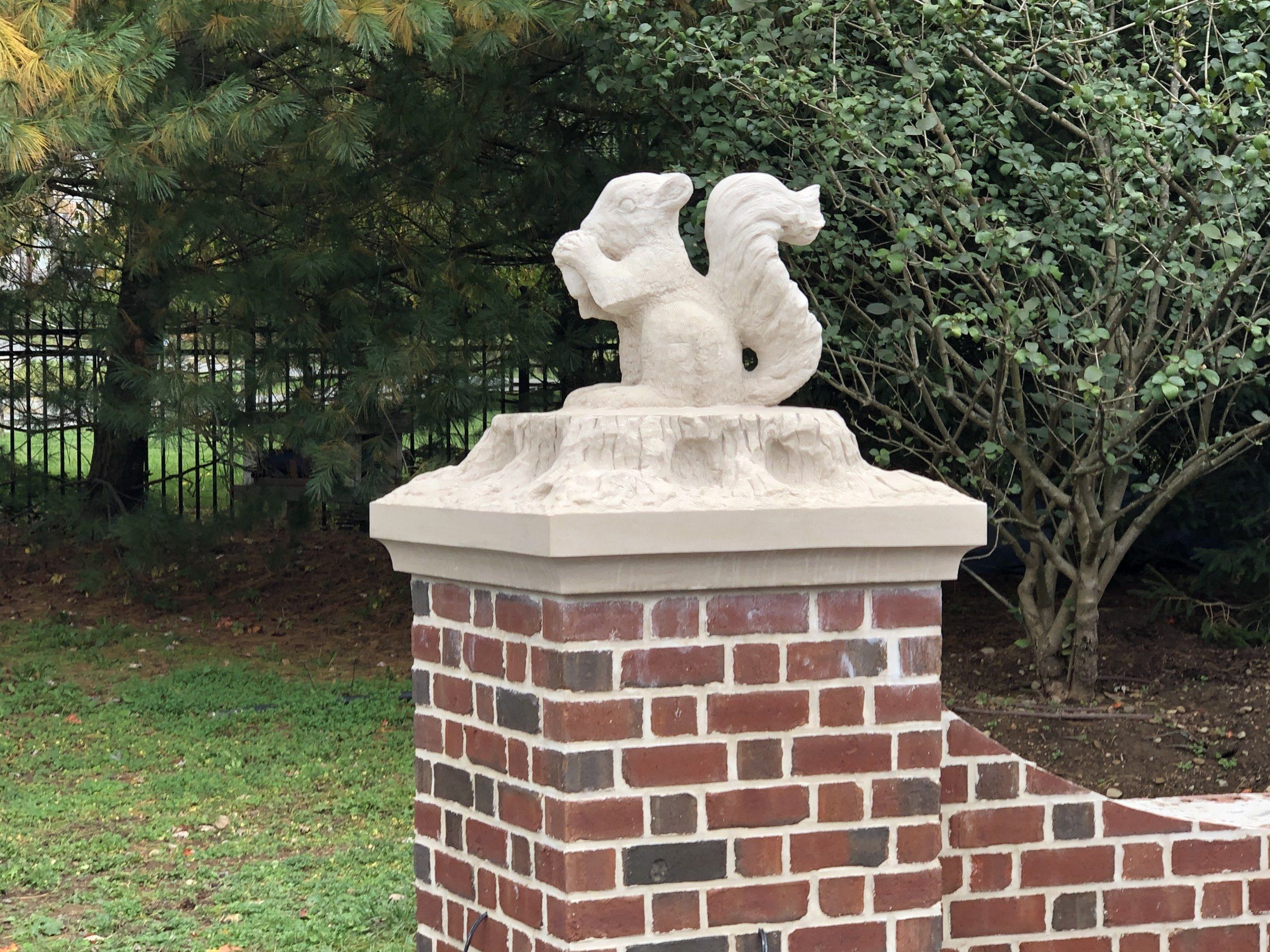 The squirrel on the tree stump, on its pier against dark evergreens and a shrub.