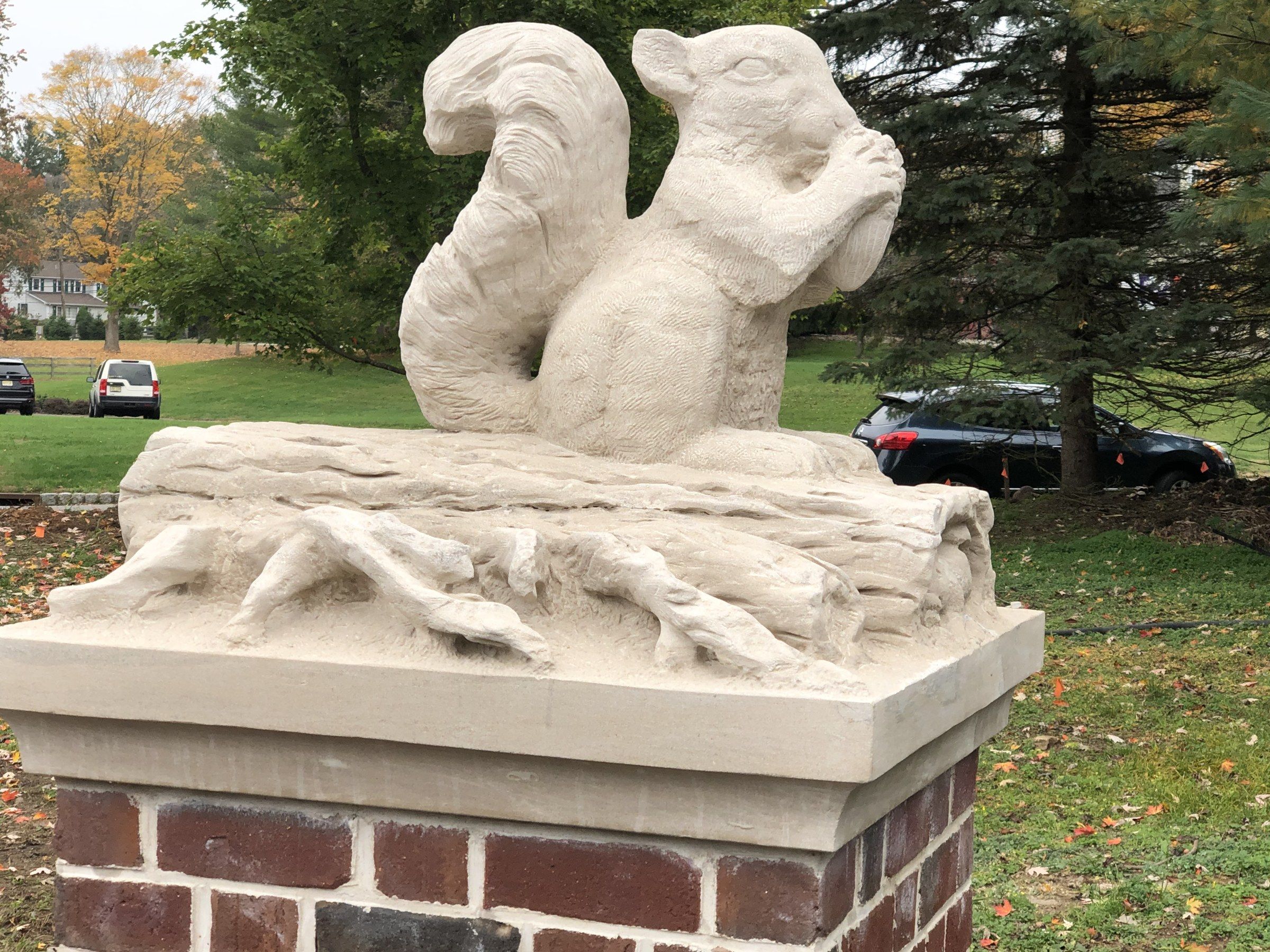 A limestone squirrel with its paws raised to its mouth and its tail curled up behind it, perched on a carved log with spreading roots on top of a limestone cap over a brick pier, with a lawn, trees and parked cars beyond.