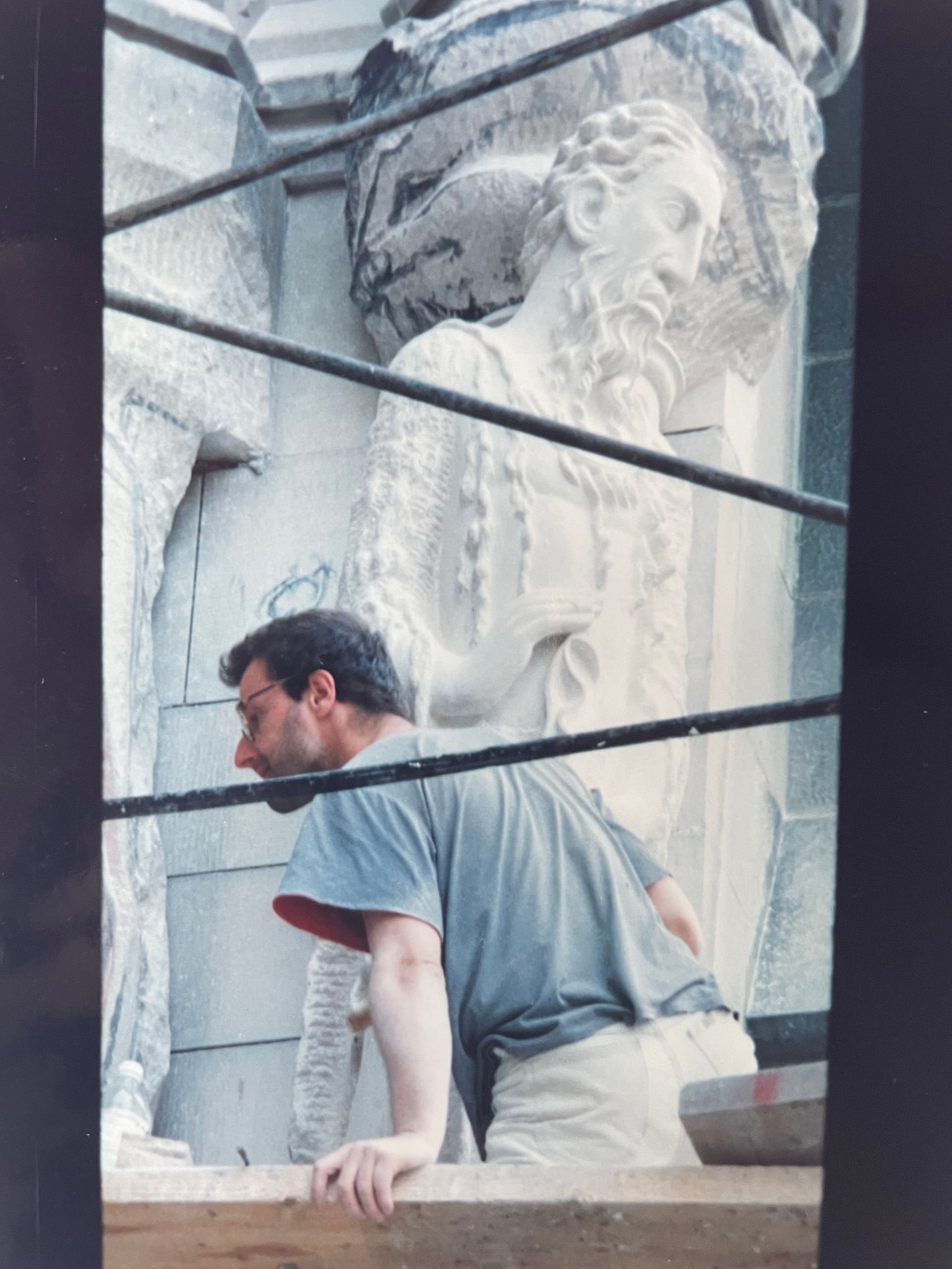 Jean-Claude Marchionni in profile, working at the Cathedral of St. John the Divine on a colossal bearded prophet figure, viewed through scaffolding bars.