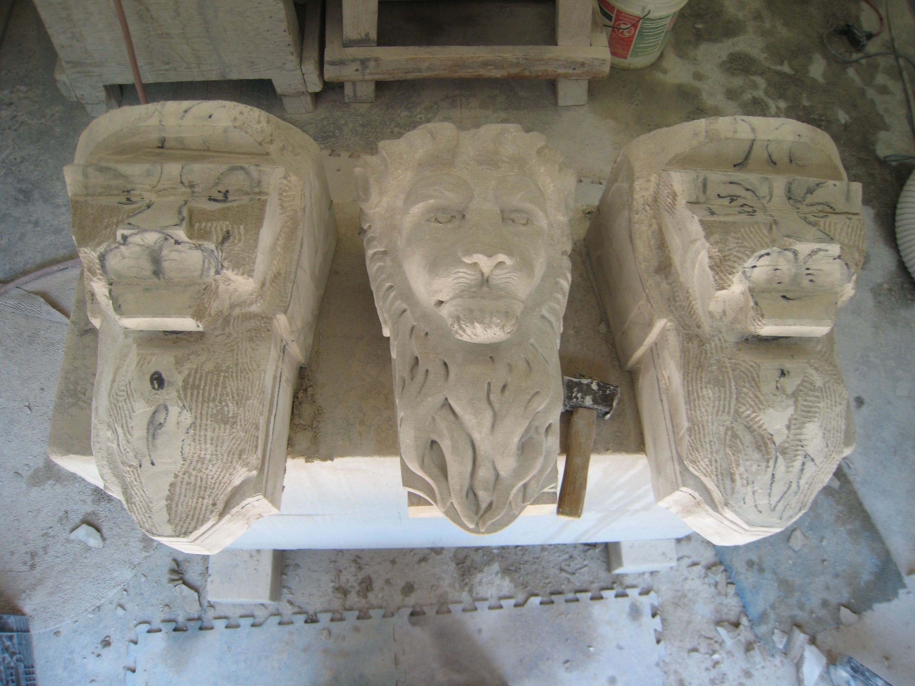 Three carved limestone lion-head corbels side by side on a workshop table, seen from above: the center one finished, with a full curling mane and a calm face, the two others still roughed out, with charcoal lines marking the features to carve.