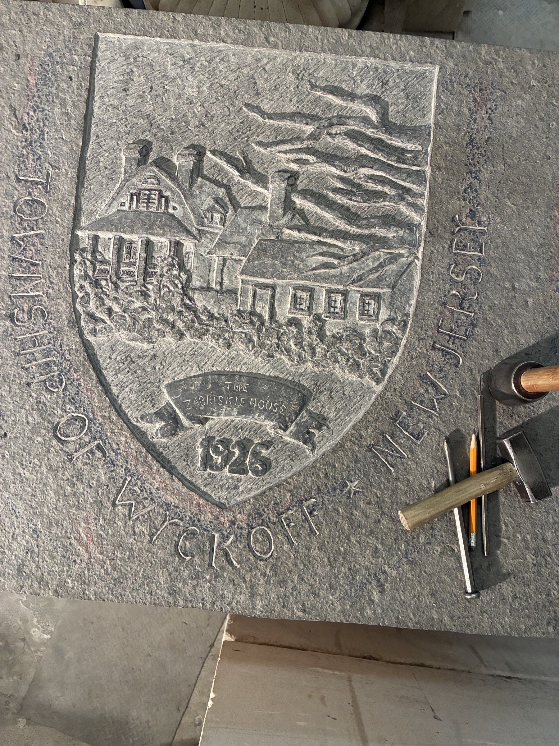 Close-up of the Township of Wyckoff coat of arms, carved in stone in March 2026: “TOWNSHIP OF WYCKOFF NEW JERSEY” around a shield with a colonial Dutch house, a tree, and the year 1926 — the start of the township's centennial, 1926–2026.