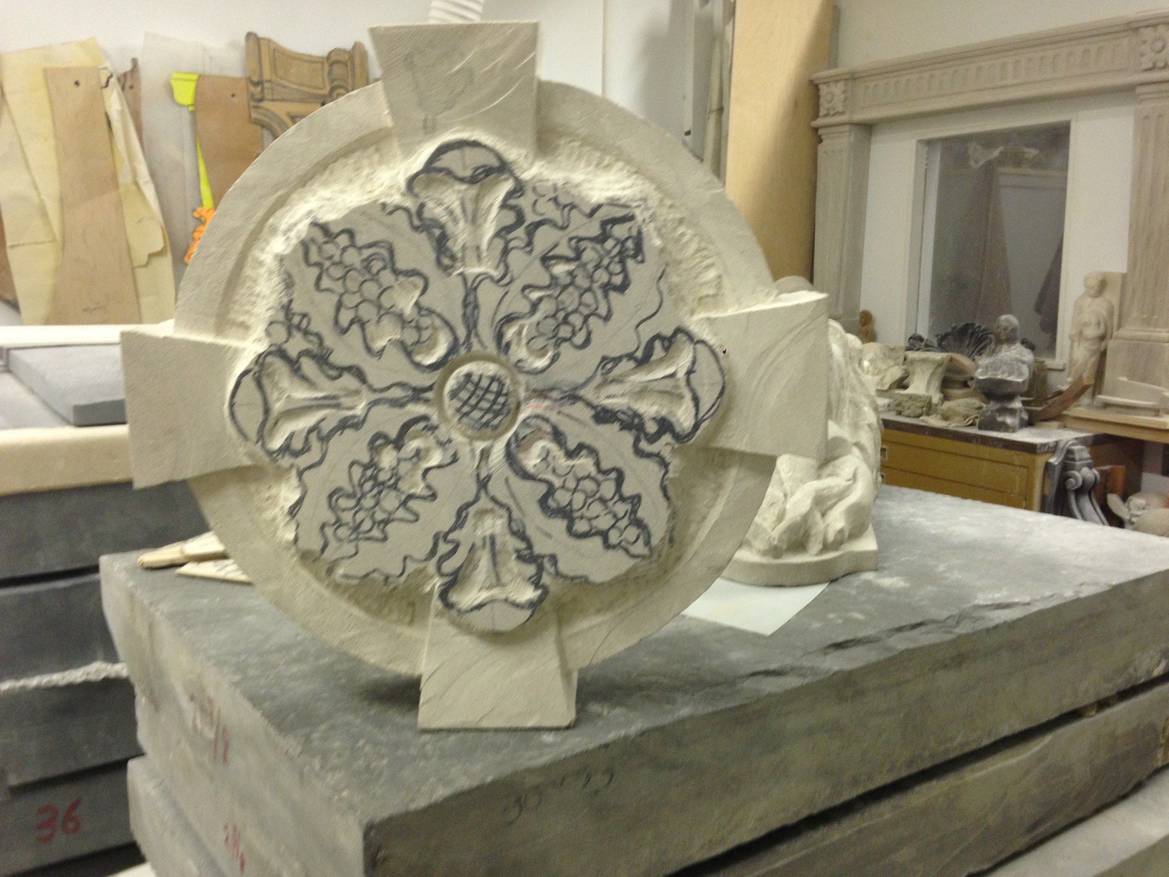 A limestone rosette standing on a stack of slabs in the studio: a round frame with four tapering arms, carved with four leafy lobes around a central boss, guide lines still drawn on the stone.