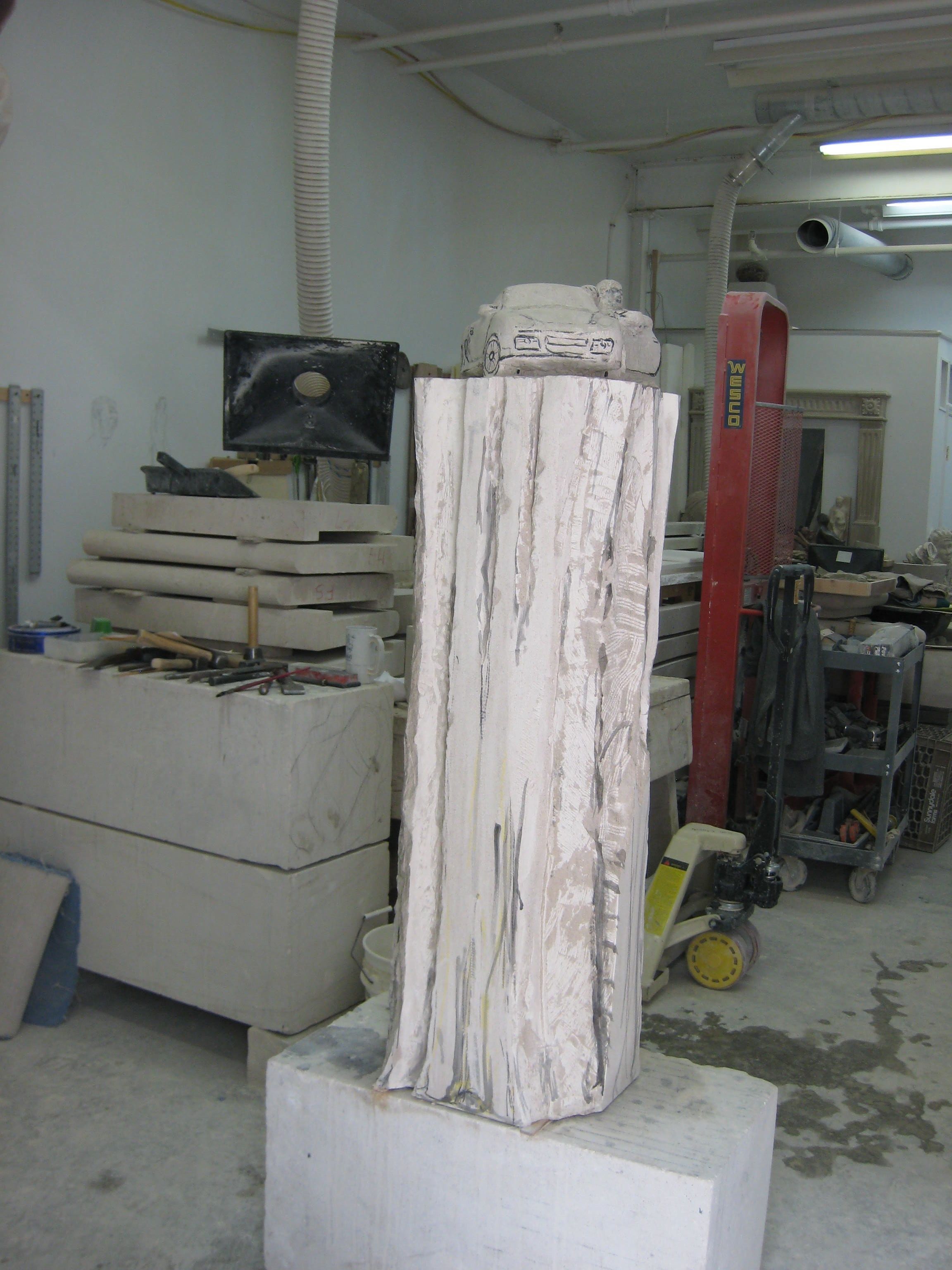 Early stage of the stone sports car — the silhouette of the vehicle blocked out at the top of a tall, roughly hewn limestone pedestal.