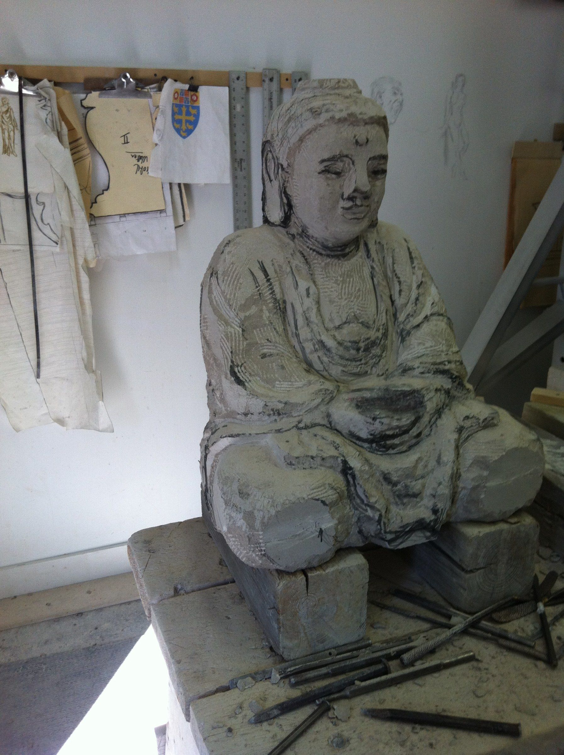 The seated Buddha roughed out on a workbench in the studio, its robe and crossed legs marked with charcoal lines, chisels lying around the base.