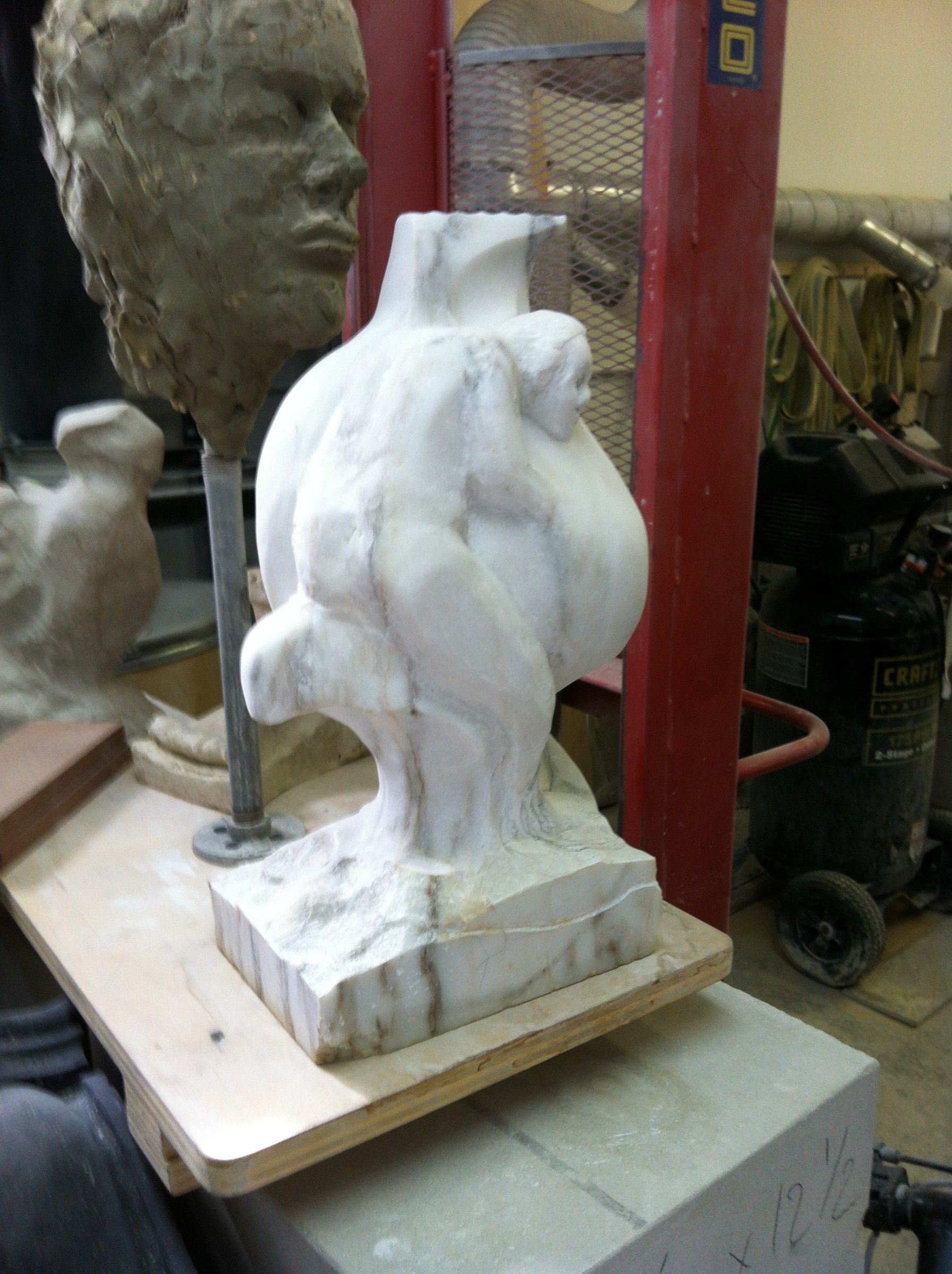 The marble at an early stage on a board in the studio: a rounded mass with a figure beginning to appear on its side, beside a clay head on a stand.