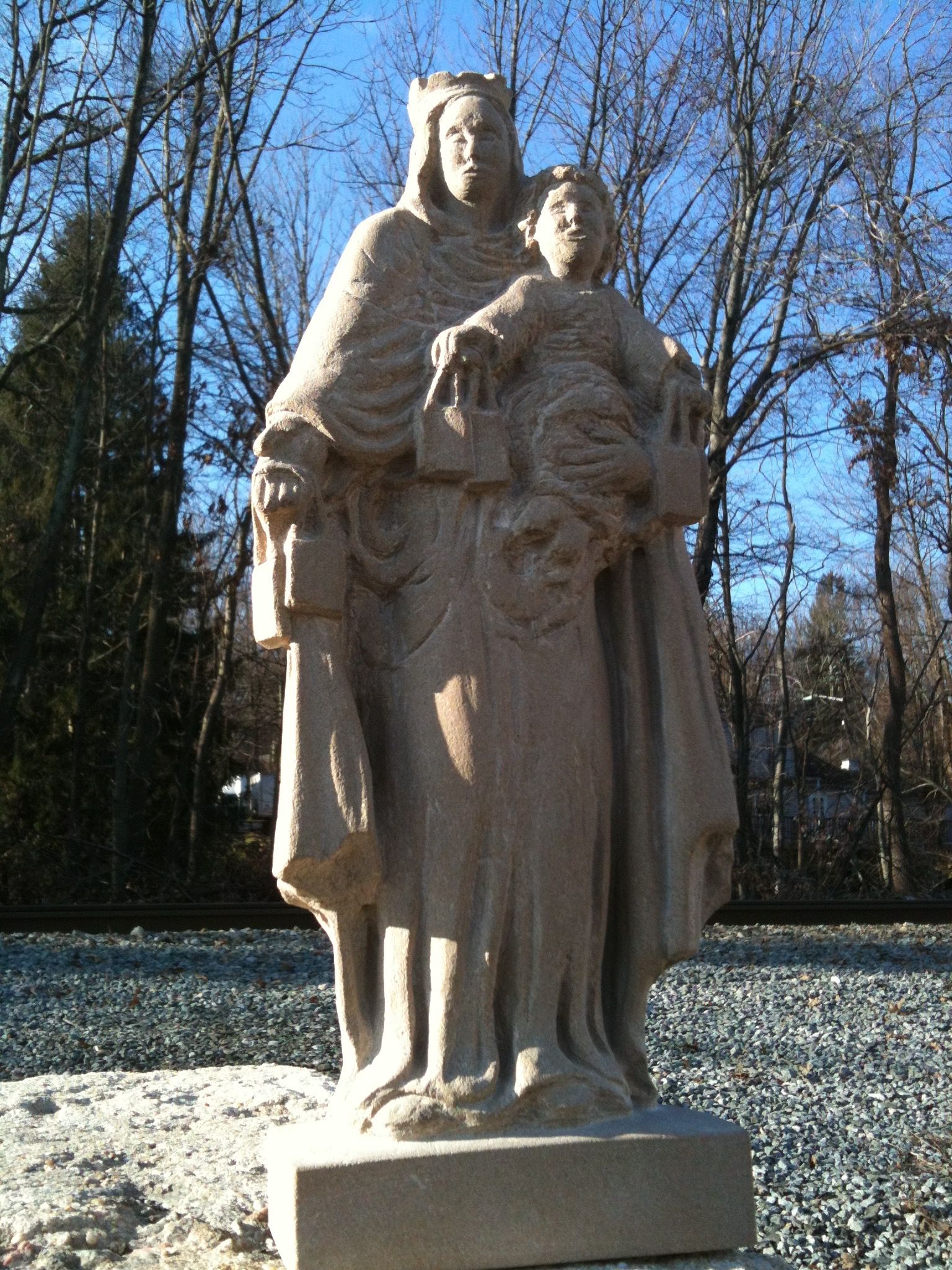 Carved limestone statue of the Madonna and Child standing outdoors against bare winter trees and a blue sky, carved by Jean-Claude Marchionni.