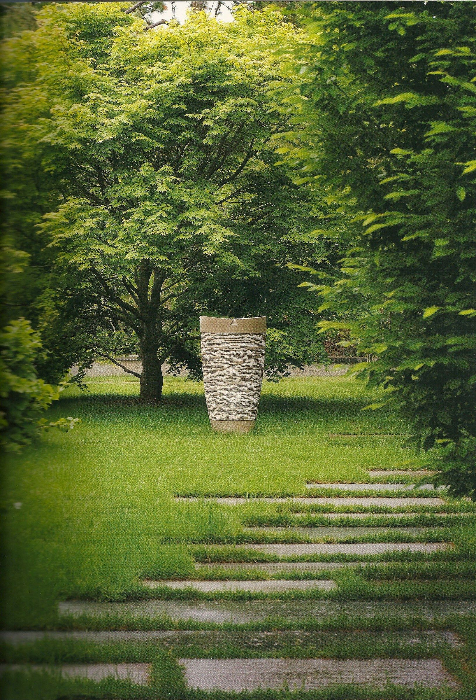 A tapering limestone stele standing at the end of a lawn path of stone stepping slabs, beneath a spreading tree and between tall hedges.