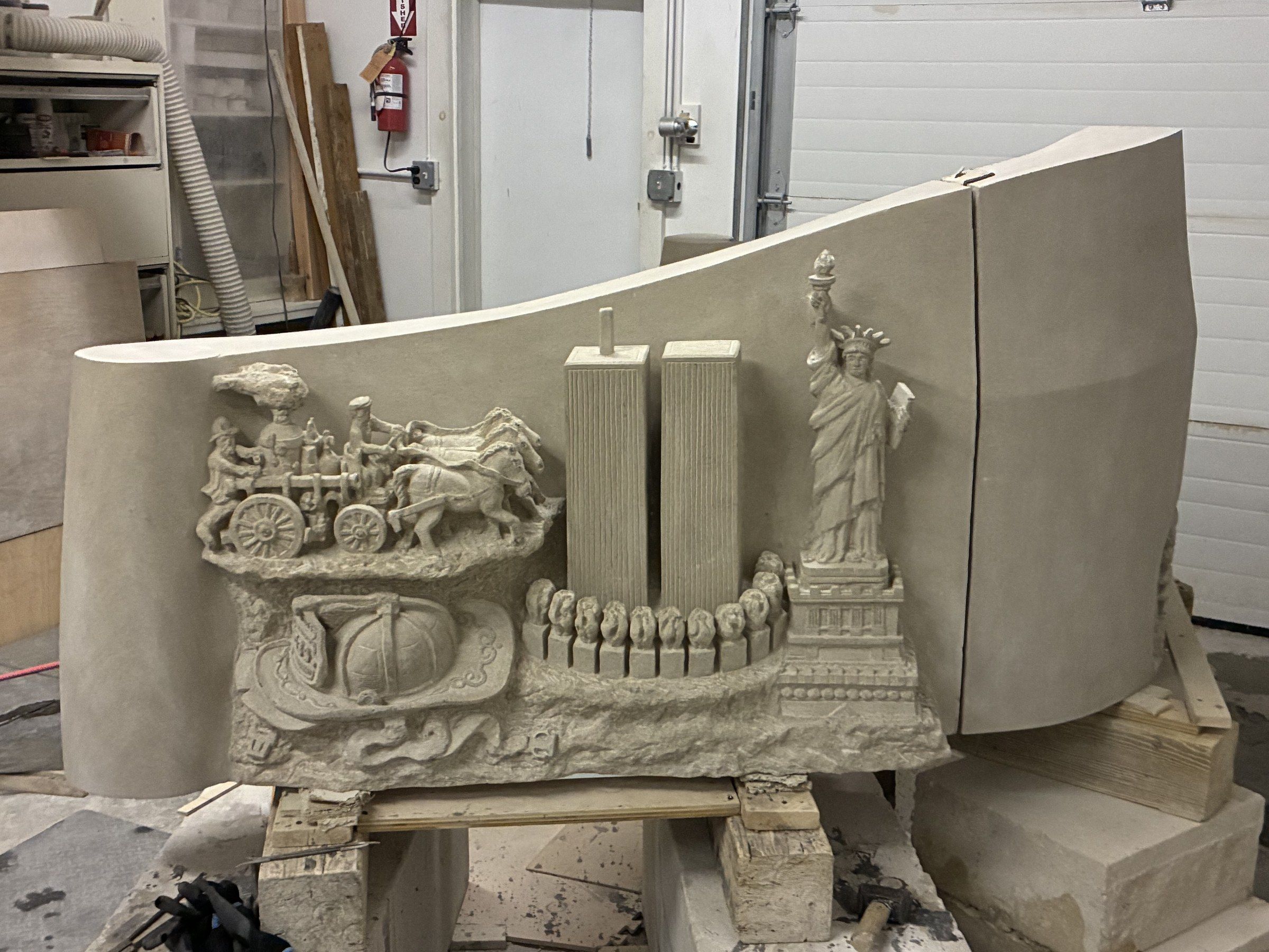 The 9/11 Memorial in Jean-Claude Marchionni's Midland Park studio, a curved three-block limestone relief raised on wooden trestles: at the centre, the Twin Towers rise straight above a row of small standing figures; at the left, a 19th-century horse-drawn fire engine with a fireman's helmet draped in the American flag at its base; at the right, the Statue of Liberty on her pedestal, torch raised. The whole piece unfolds in a single flowing wave.