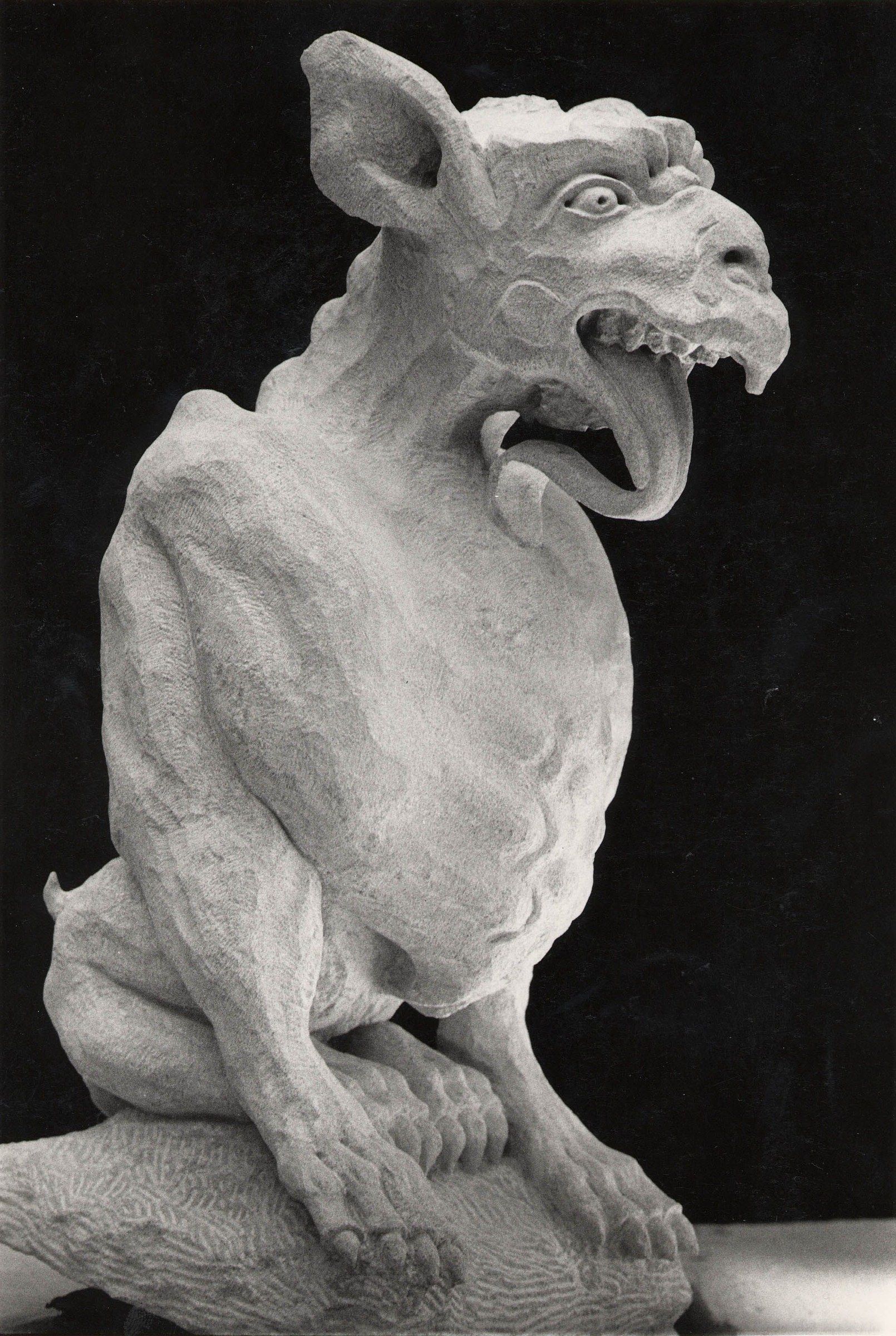 Black-and-white photograph of a carved limestone chimera against a black backdrop: a lean, seated creature with tall pointed ears, a hooked, beaklike snout, and open jaws lined with sharp teeth, its long tongue curling out and down; ridged skin folds over the shoulders and flanks, and clawed forepaws grip a rough, textured base.