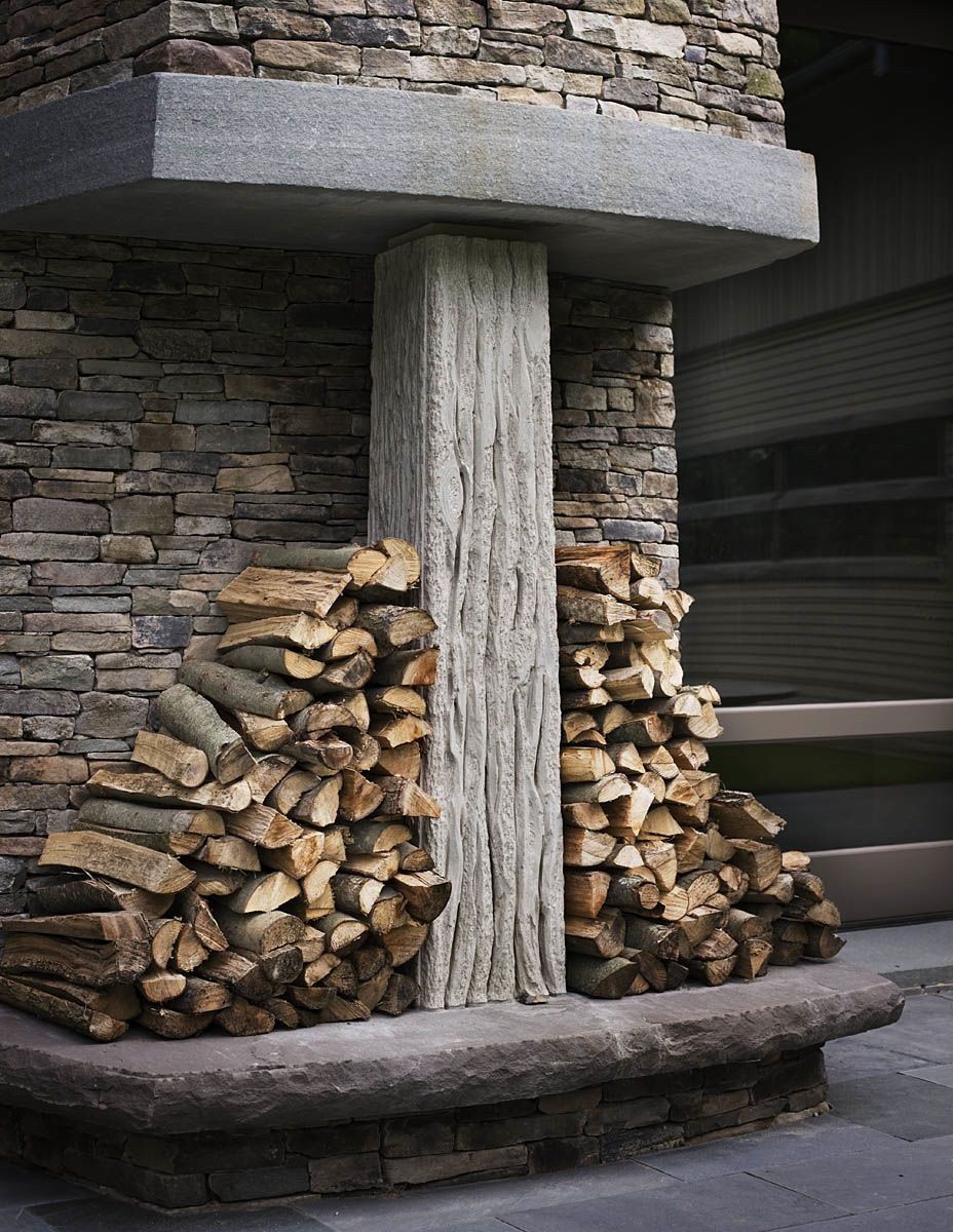 Close-up of the fireplace base: the carved limestone column with deep vertical fluting that reads as bark, flanked by two symmetrical stacks of split firewood against walls of stacked stone.