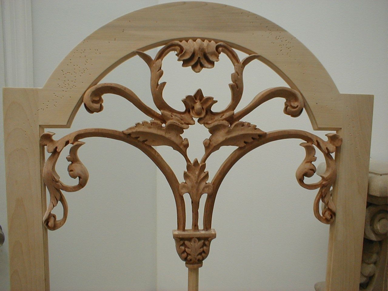 An unfinished carved wood panel set in a pale round-arched frame: openwork scrolls of acanthus rise in symmetrical arcs from a small carved cup at the base, meeting at a central flower and a crowning palmette, the spaces between left open.